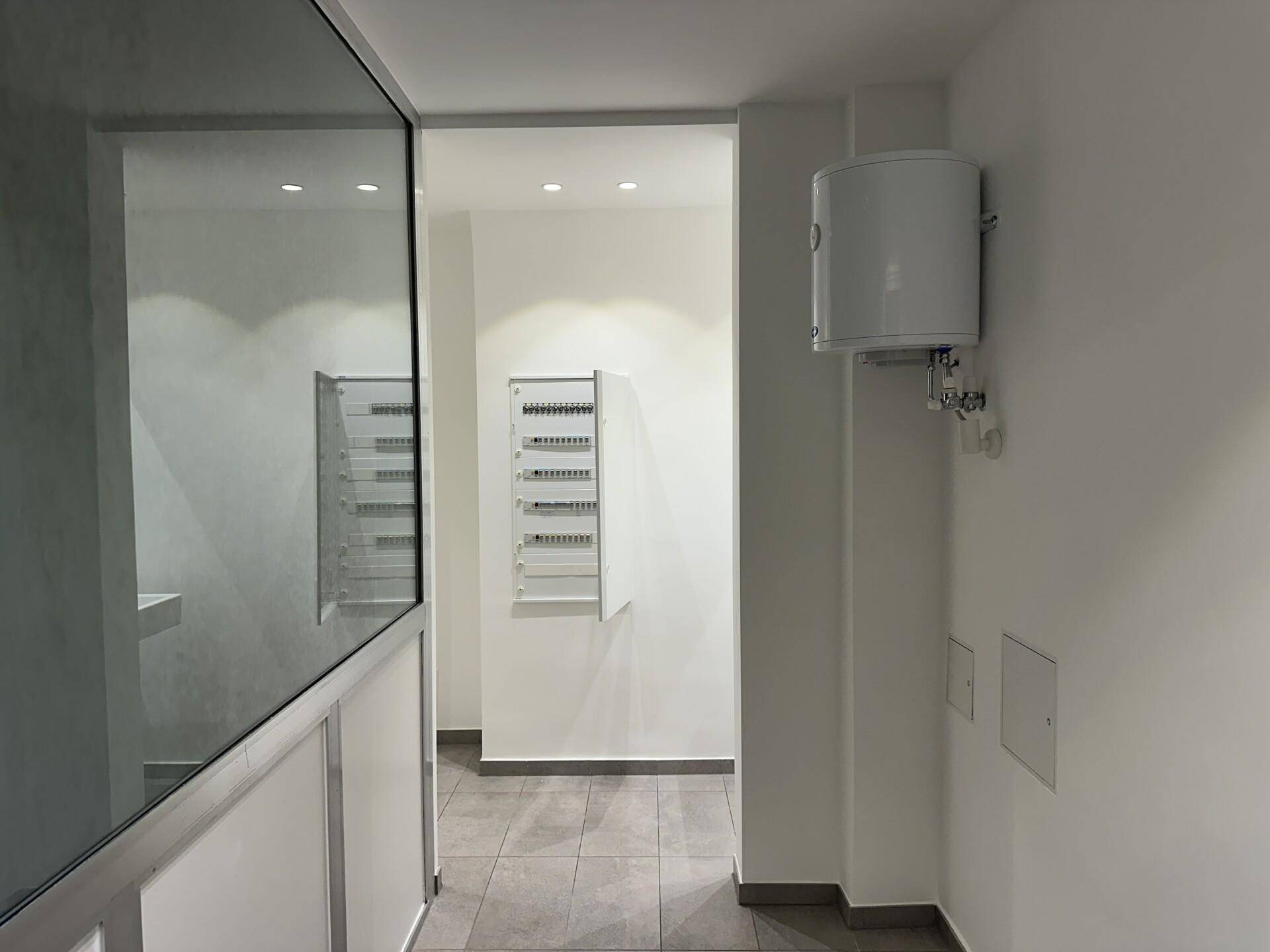 Funktionaler Bereich mit grauem Fliesenboden, weiß gestrichenen Wänden und sichtbarem Warmwasserbereiter.