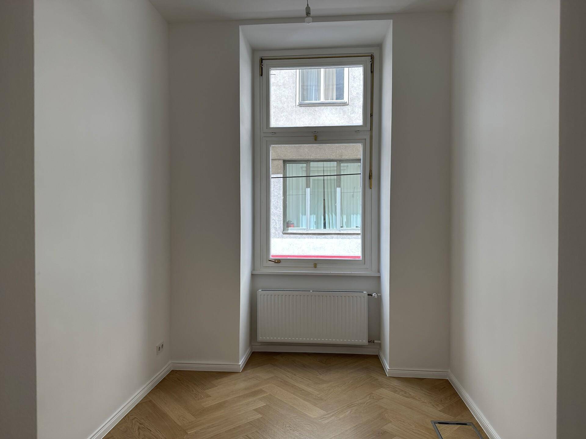 Helles Zimmer mit hohem Fenster, Fischgrätparkett und weiß gestrichenen Wänden, bietet vielseitige Nutzungsmöglichkeiten.