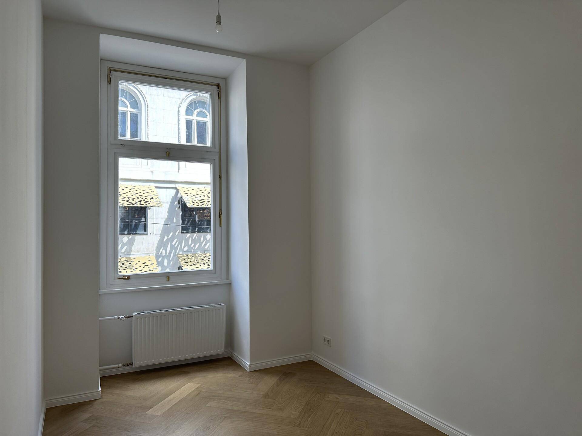 Helles Zimmer mit hohem Fenster, Fischgrätparkett und weiß gestrichenen Wänden, bietet vielseitige Nutzungsmöglichkeiten.