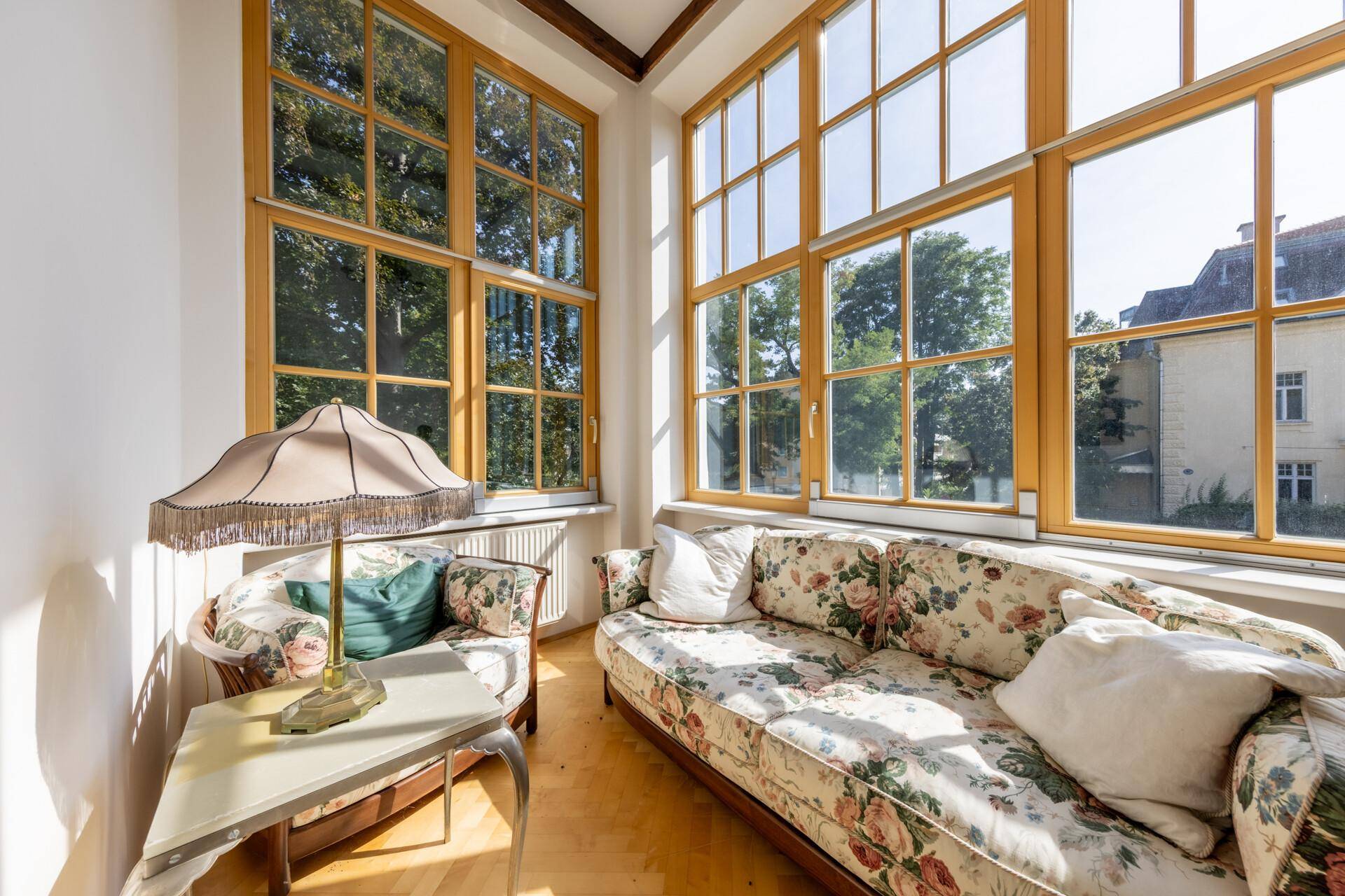 Helles Wohnzimmer mit großen Fenstern, floralem Sofa und Holzboden.