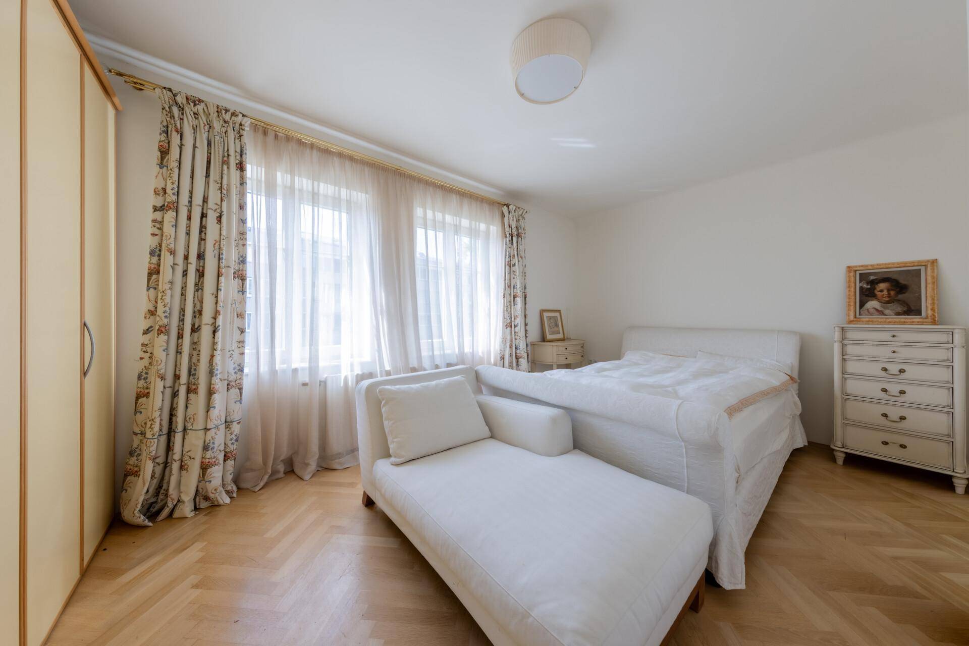 Helles Schlafzimmer mit Parkettboden, weißem Bett und floralen Vorhängen.
