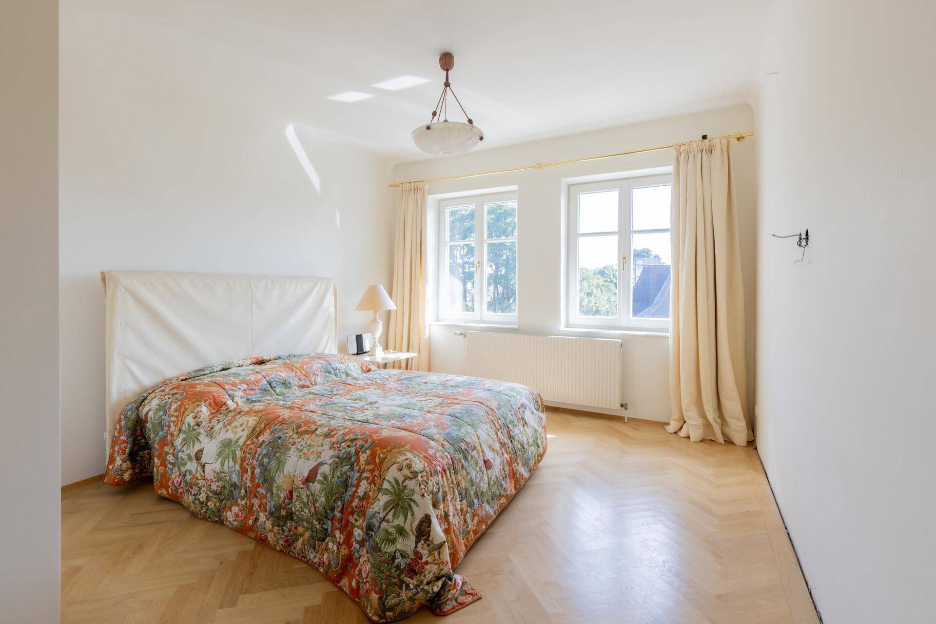 Schlafzimmer mit Parkettboden, farbenfroher Bettdecke und großen Fenstern.