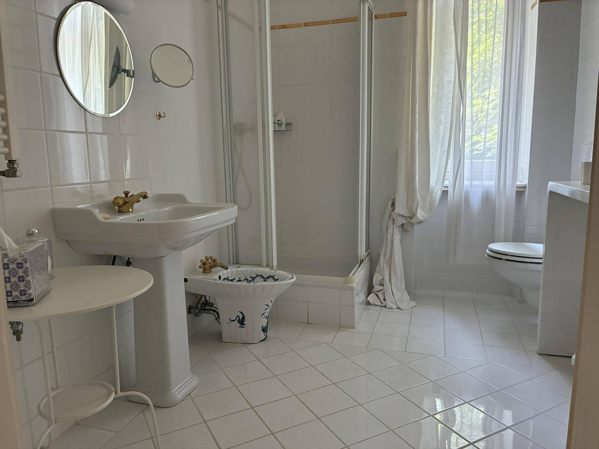 Badezimmer mit weißem Fliesenboden, Dusche, Bidet und Fenster für Tageslicht.