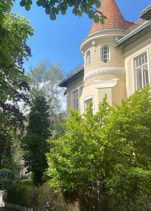 Historisches Gebäude mit markantem Turm und grüner Umgebung unter blauem Himmel.