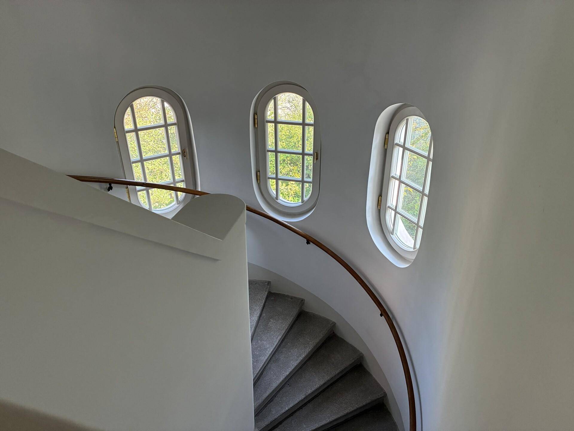 Helles Treppenhaus mit geschwungener Steintreppe und drei ovalen Fenstern.