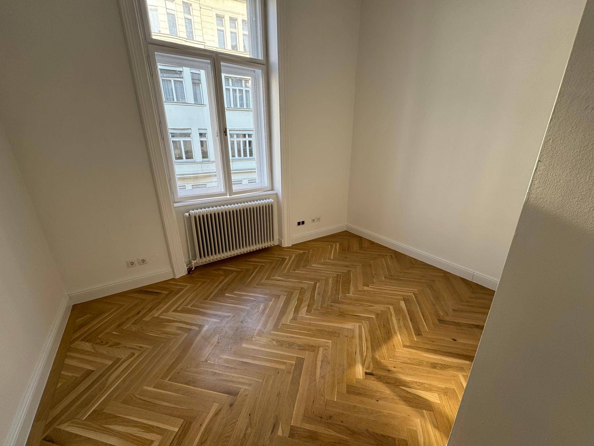 Helles Eckzimmer mit Fischgrätparkett, weißen Wänden und einem großen Fenster mit Heizkörper.