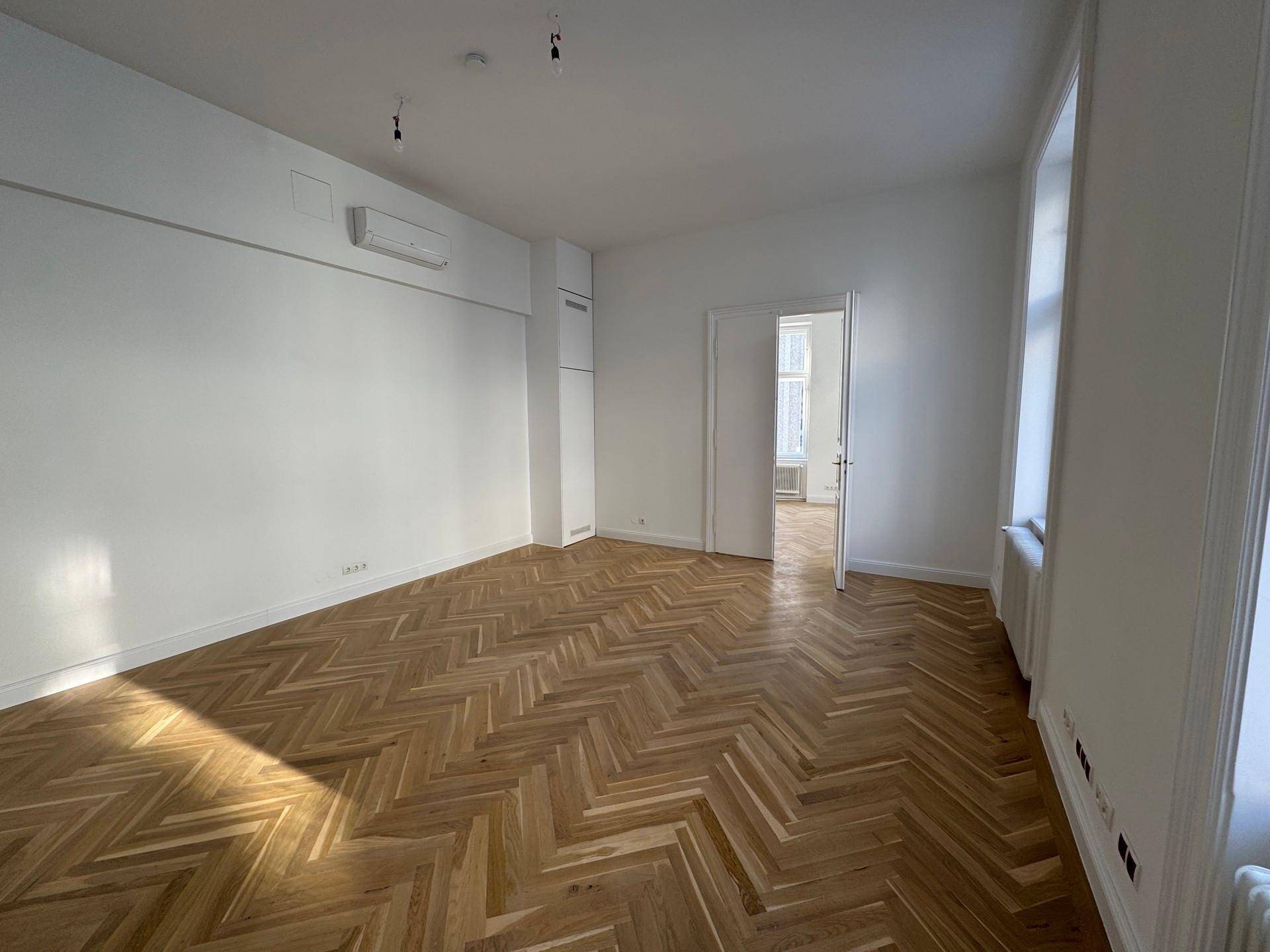 Geräumiges Zimmer mit Fischgrätparkett, weißen Wänden, Klimaanlage und Zugang zu einem weiteren Raum.