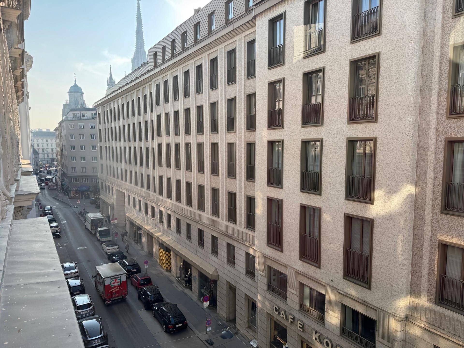 Blick auf eine städtische Straße mit Gebäuden, parkenden Autos und einer Kirchturmspitze in der Ferne.