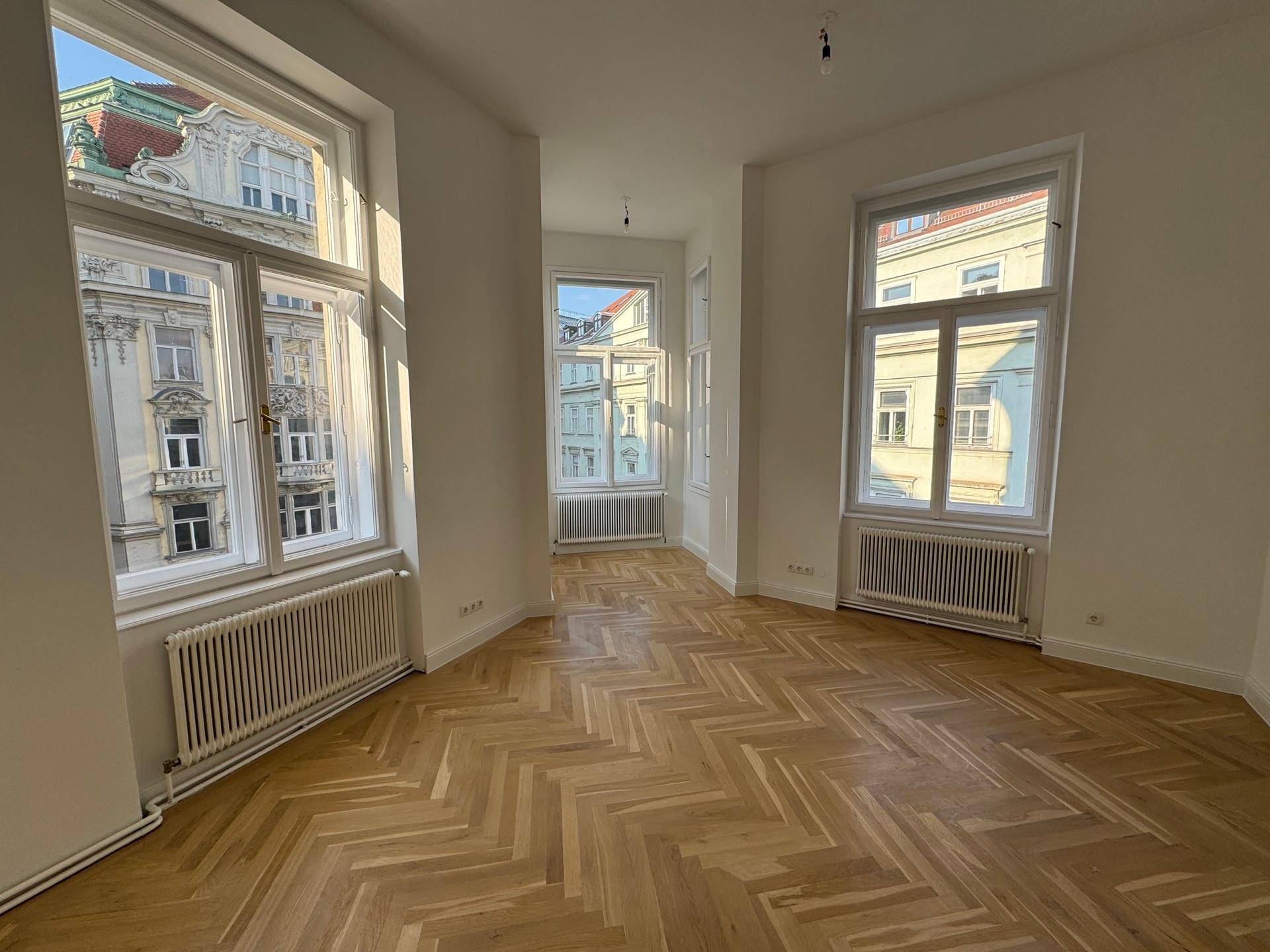 Heller Wohnraum mit Fischgrätparkett, hohen Decken und mehreren großen Fenstern mit Blick auf historische Gebäude.
