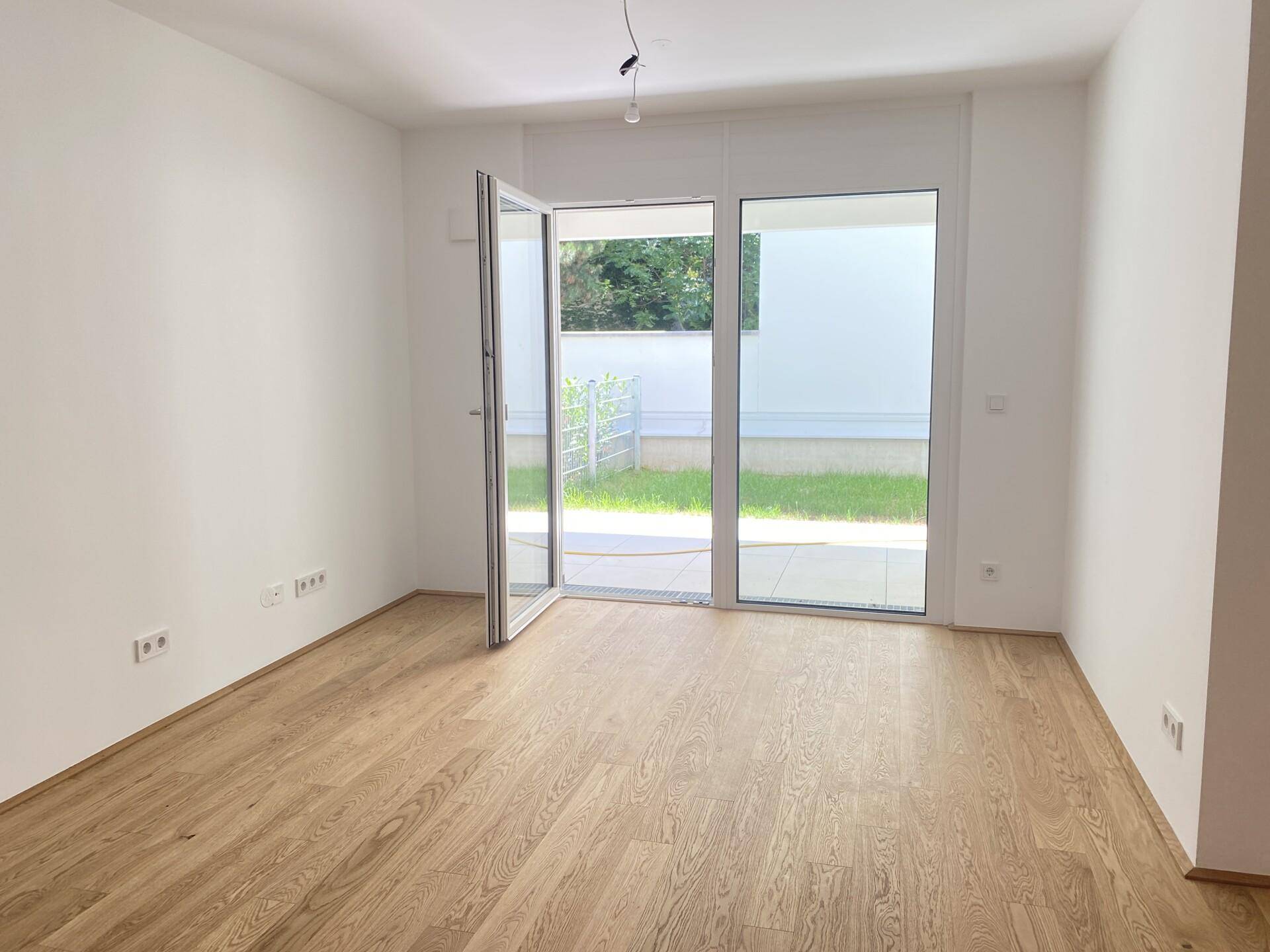Heller, leerer Raum mit hellem Holzparkett und großen Glastüren, die zum Garten führen.