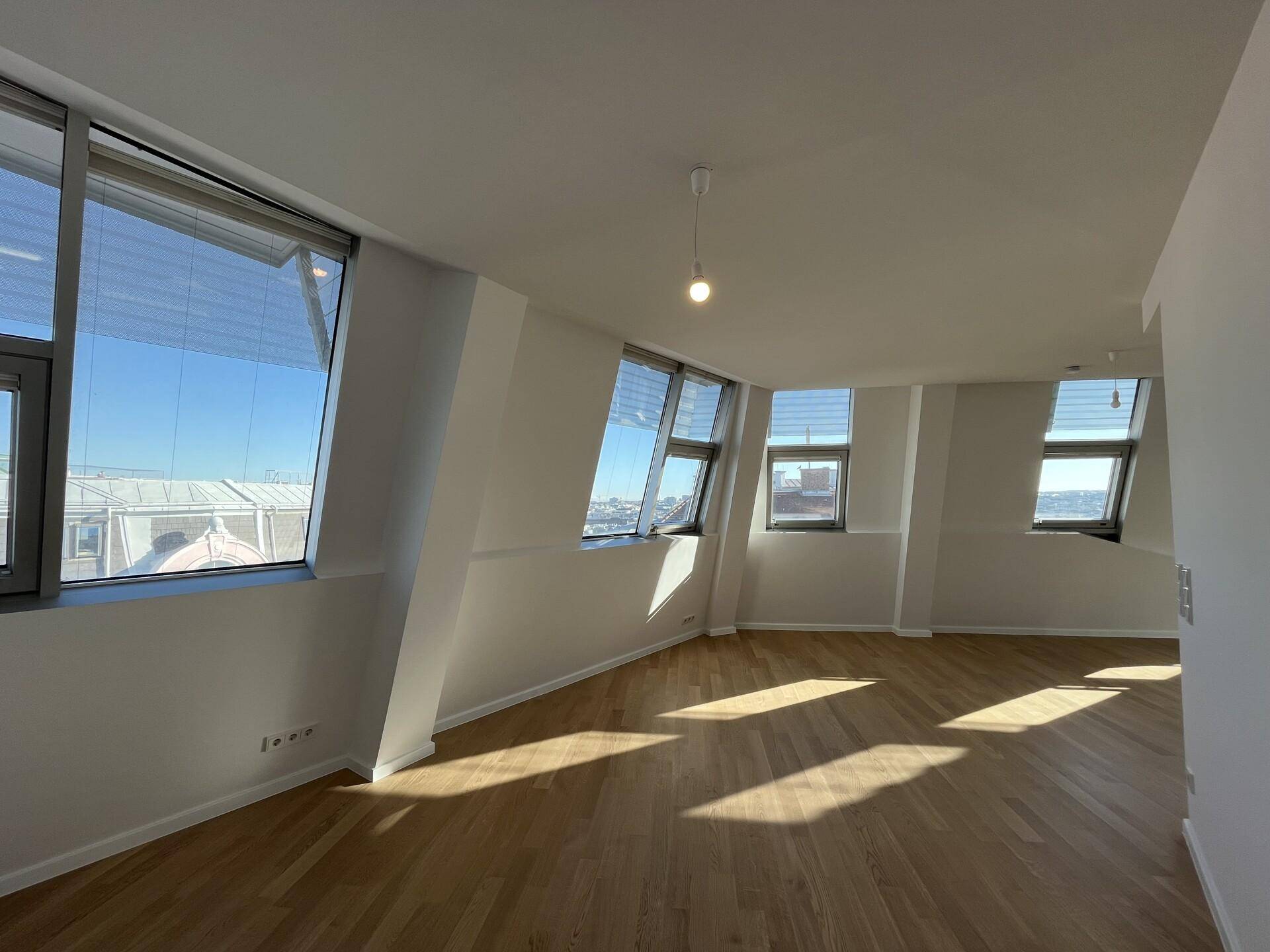 Heller Raum mit Holzboden und vielen Fenstern, die einen weiten Blick über die Stadt bieten.