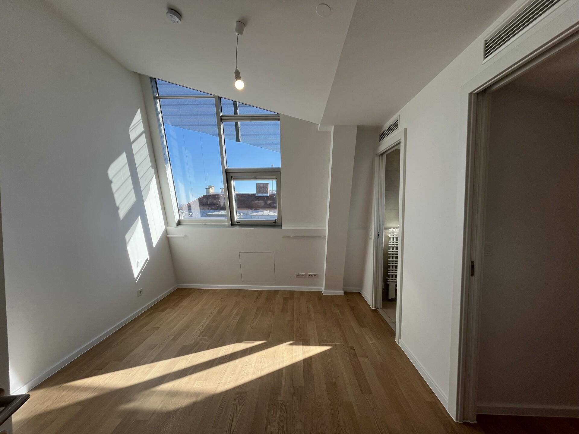 Lichtdurchflutetes Schlafzimmer mit Holzboden und schrägen Fenstern, angrenzend an ein Bad.