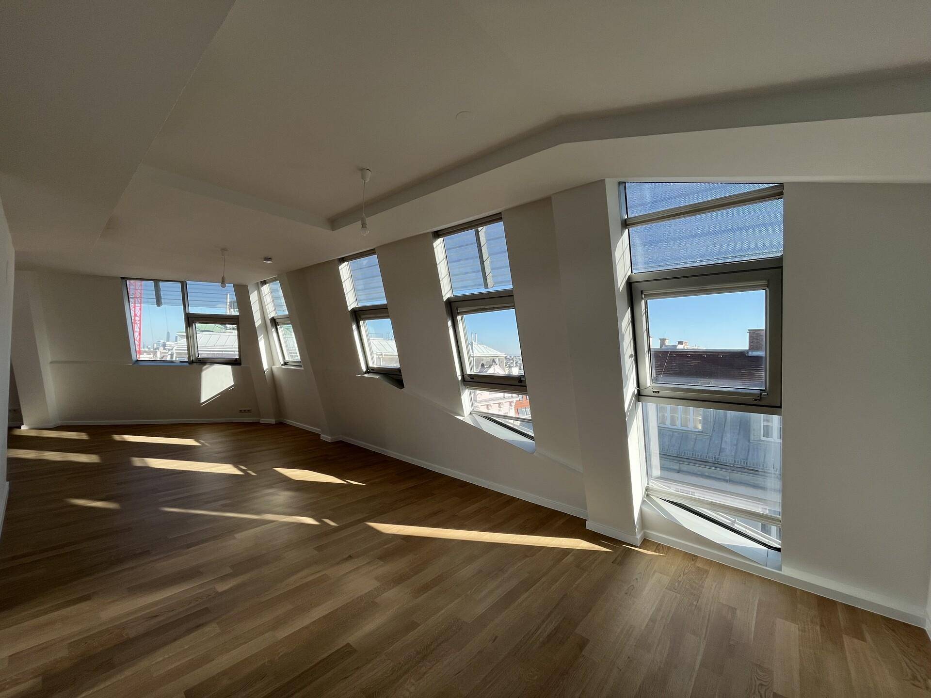 Großzügiger, heller Wohnraum mit Holzboden und vielen Fenstern, die viel Licht hereinlassen.
