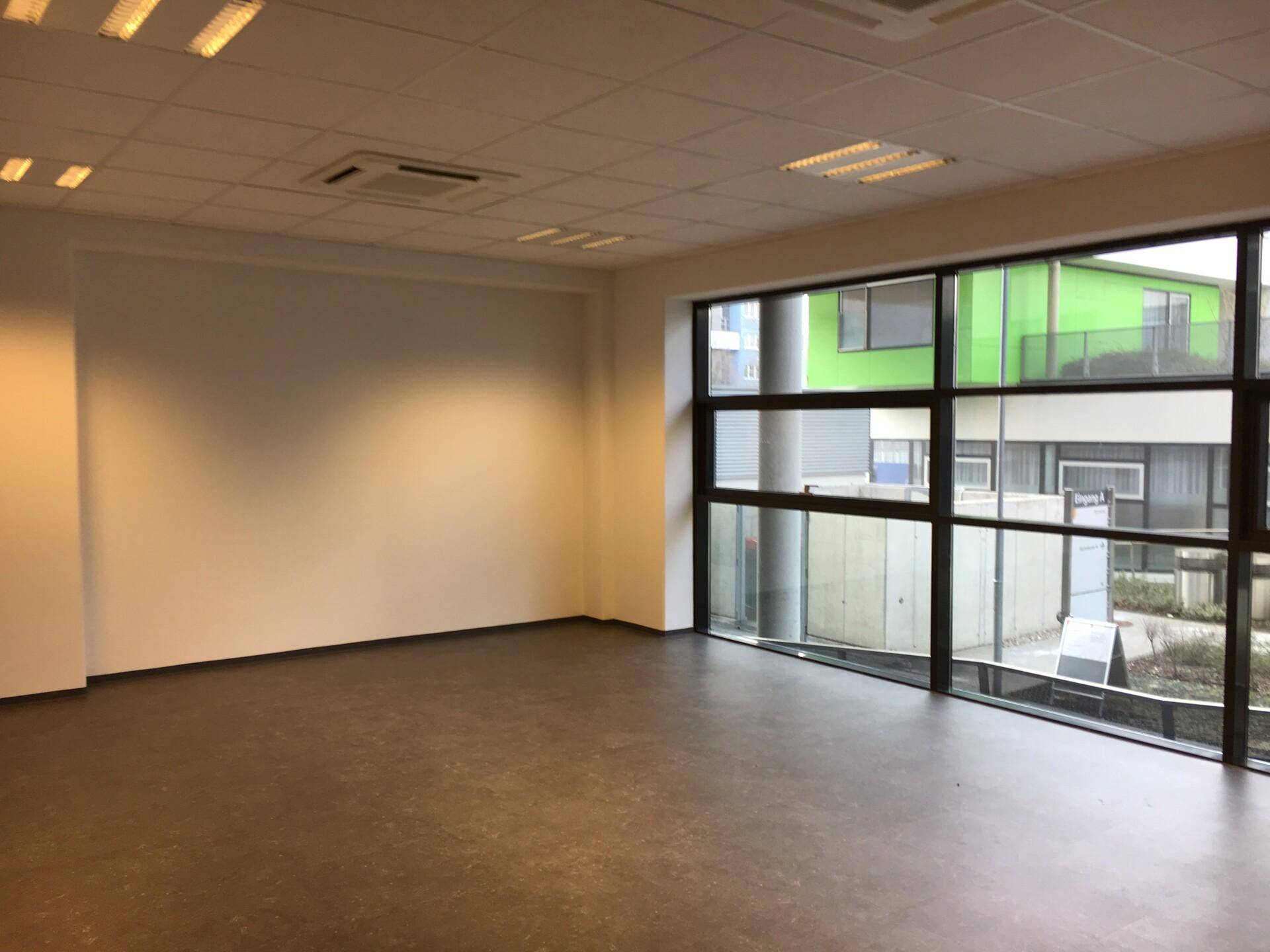 Geräumiger Büroraum mit großen Fenstern, dunklem Boden und Blick auf umliegende Gebäude.
