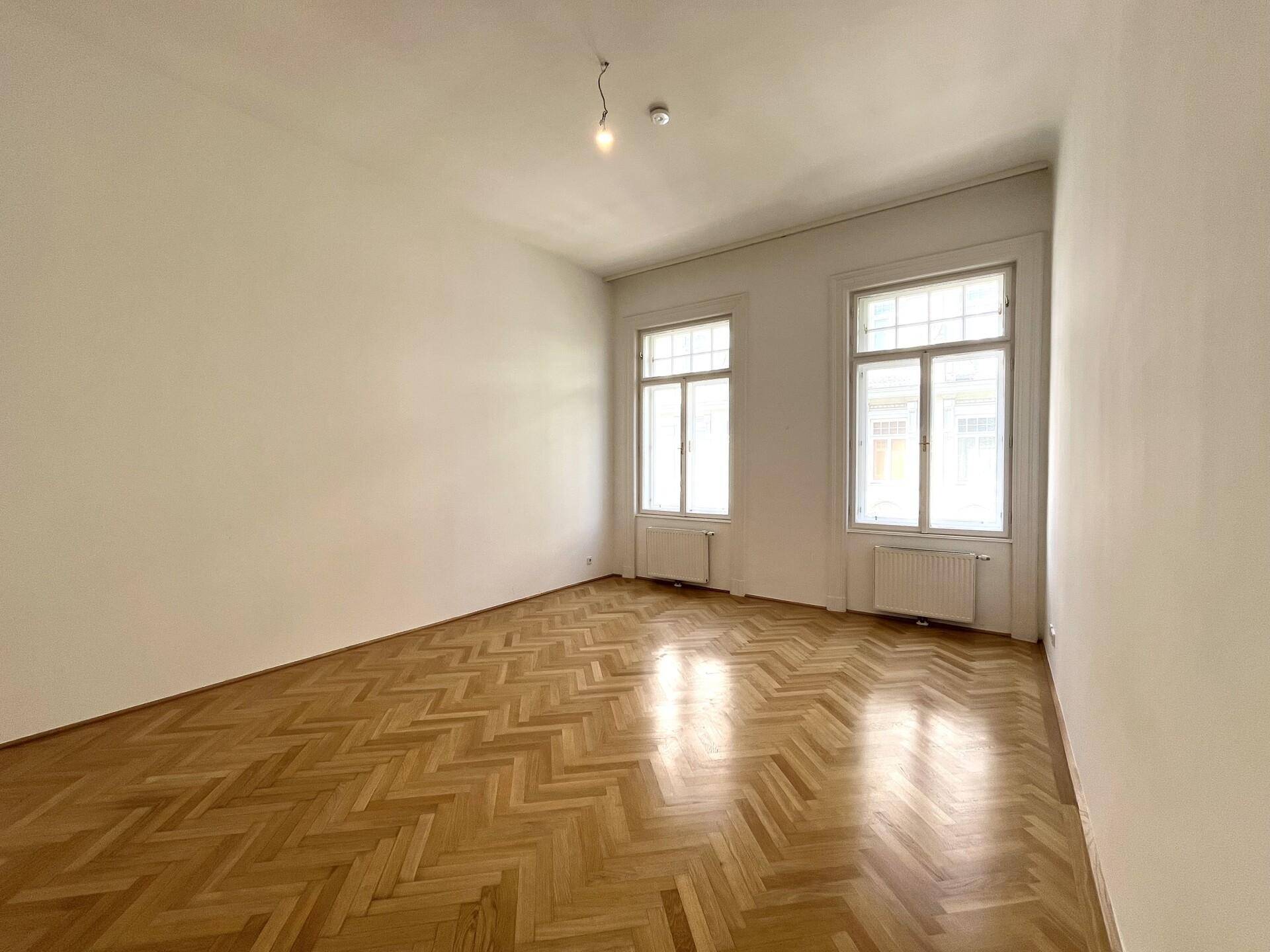Helles Schlafzimmer mit klassischem Fischgrätparkett und zwei hohen Fenstern, die viel Licht spenden.