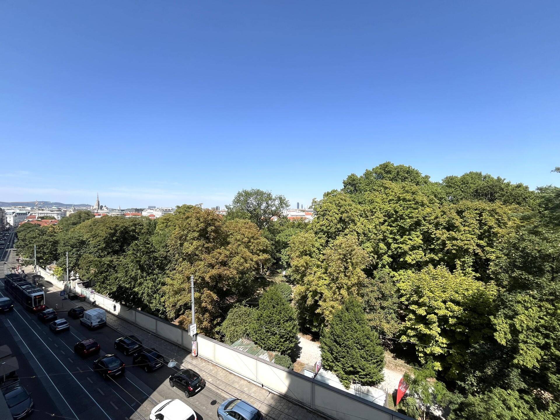 Weitläufiger Blick über eine belebte Straße und einen großen Park mit Stadtpanorama im Hintergrund.