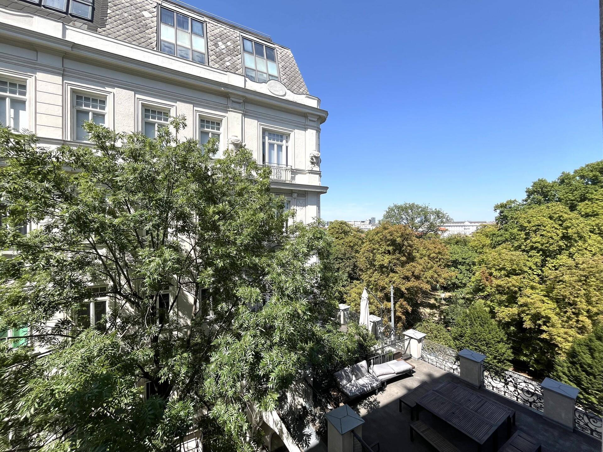 Sonniger Außenbereich mit Terrasse und Sitzgelegenheiten, umgeben von grünen Bäumen und Blick auf die Stadt.