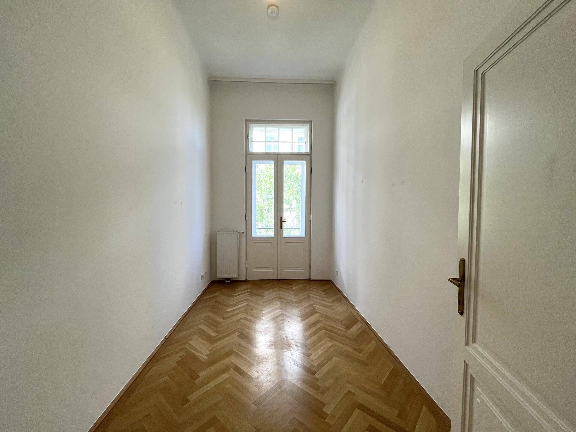 Langer, heller Flur mit Fischgrätparkett und einer Doppeltür, die nach draußen führt.