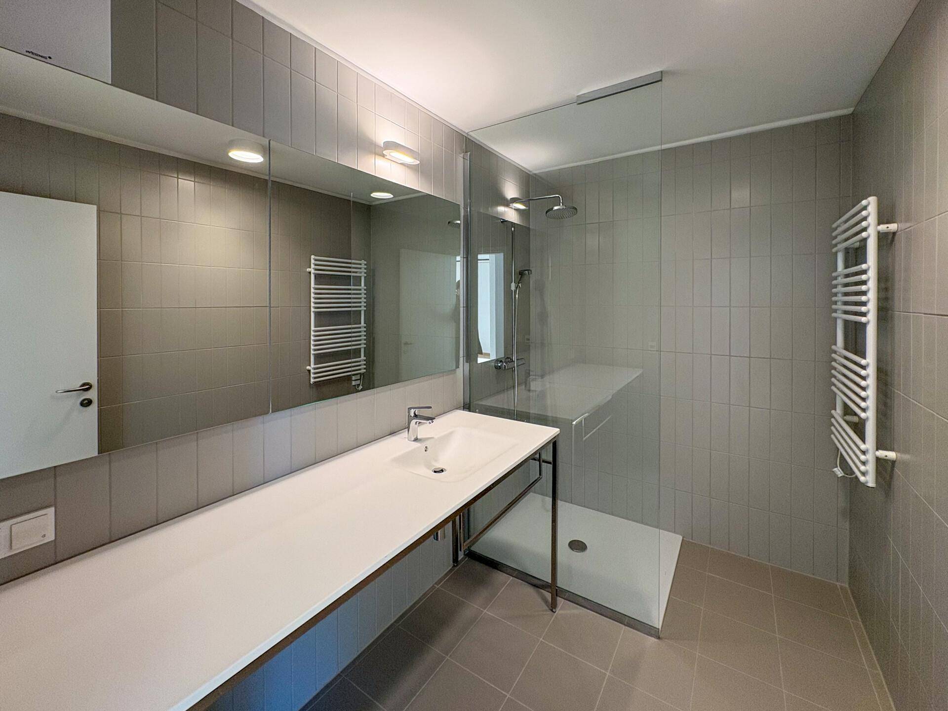 Modernes Badezimmer mit grauen Fliesen, großem Waschtisch, begehbarer Dusche und beheiztem Handtuchhalter.