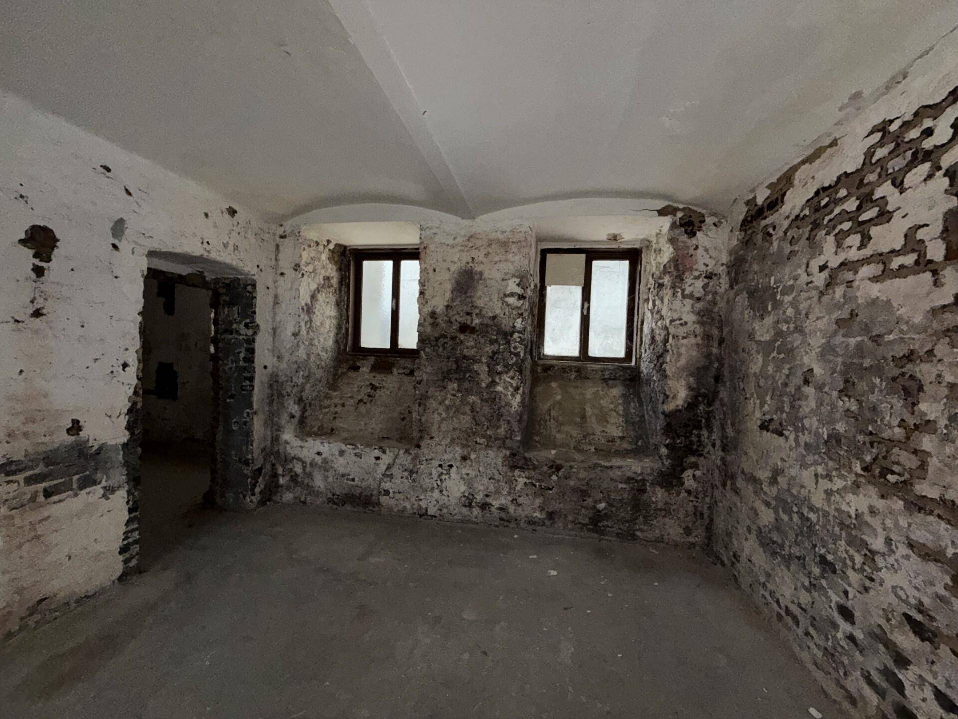 Kellerraum mit zwei kleinen Fenstern und stark abgenutzten, freigelegten Ziegelwänden.