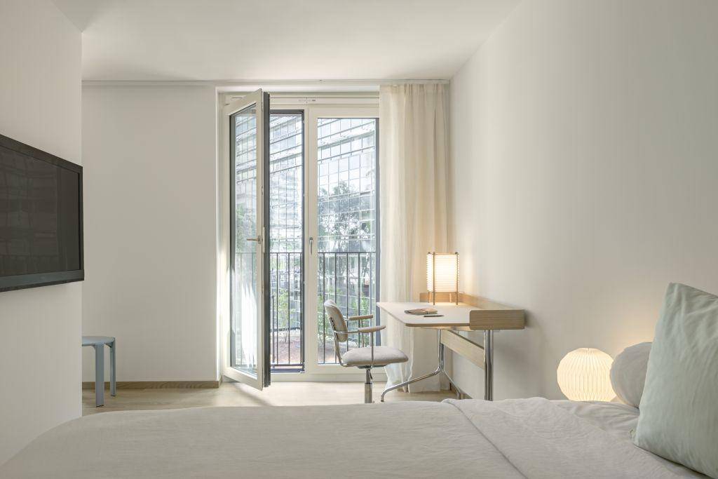 Schlafzimmer mit Bett, Schreibtisch und Zugang zu einem französischen Balkon, hell und modern.