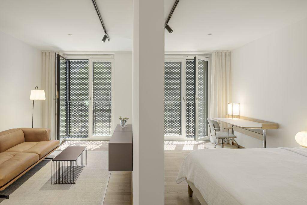 Heller Wohnbereich mit Sofa, Schreibtisch und Bett, durch eine Wand getrennt, mit Zugang zum Balkon.