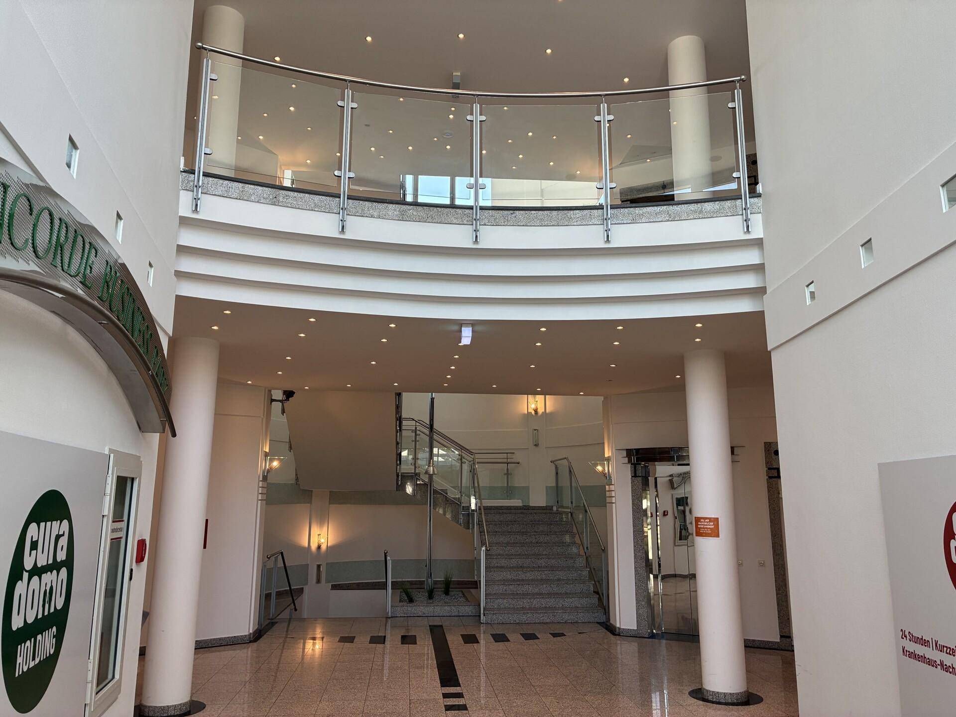Heller, mehrstöckiger Eingangsbereich mit geschwungener Treppe, Glasgeländer und vielen Deckenspots.