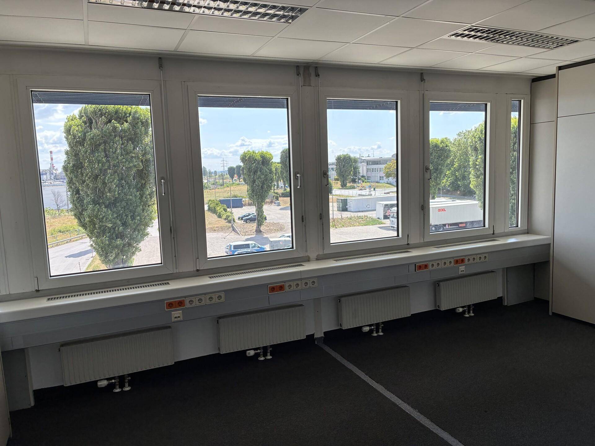 Große Fensterfront in einem Büro mit Blick auf Bäume und einen Gewerbepark, darunter Heizkörper und Steckdosen.