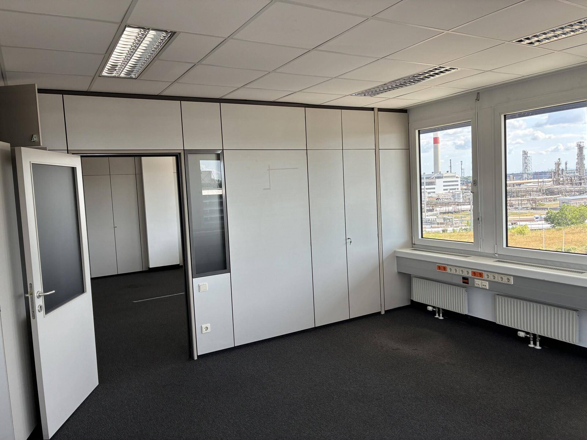 Leeres Büro mit modularen Trennwänden, einer Tür zu einem weiteren Raum und Blick auf eine Industrieanlage.