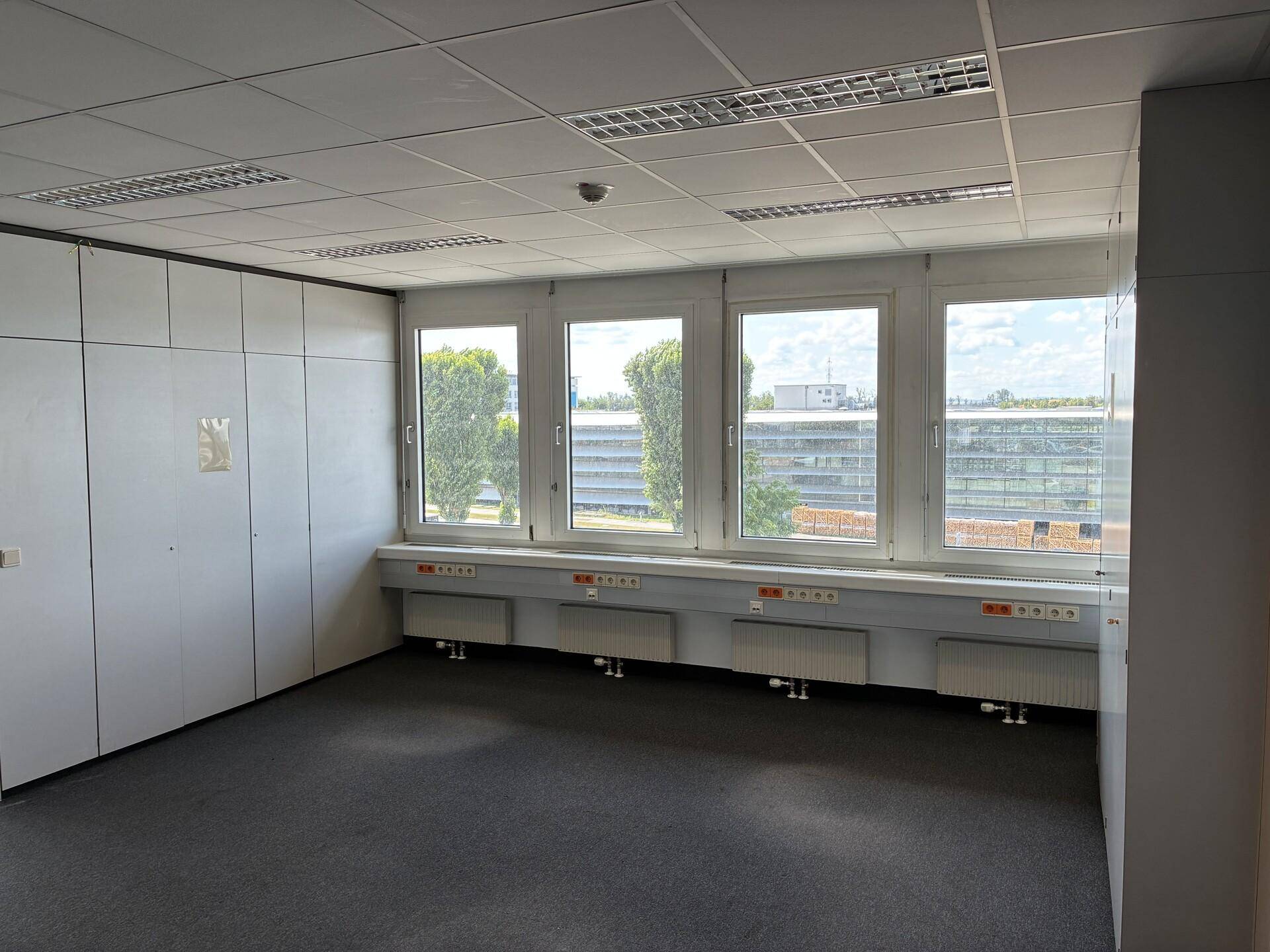 Weitläufiges Büro mit einer langen Fensterfront, modularen Wänden und Blick auf umliegende Gebäude und Bäume.