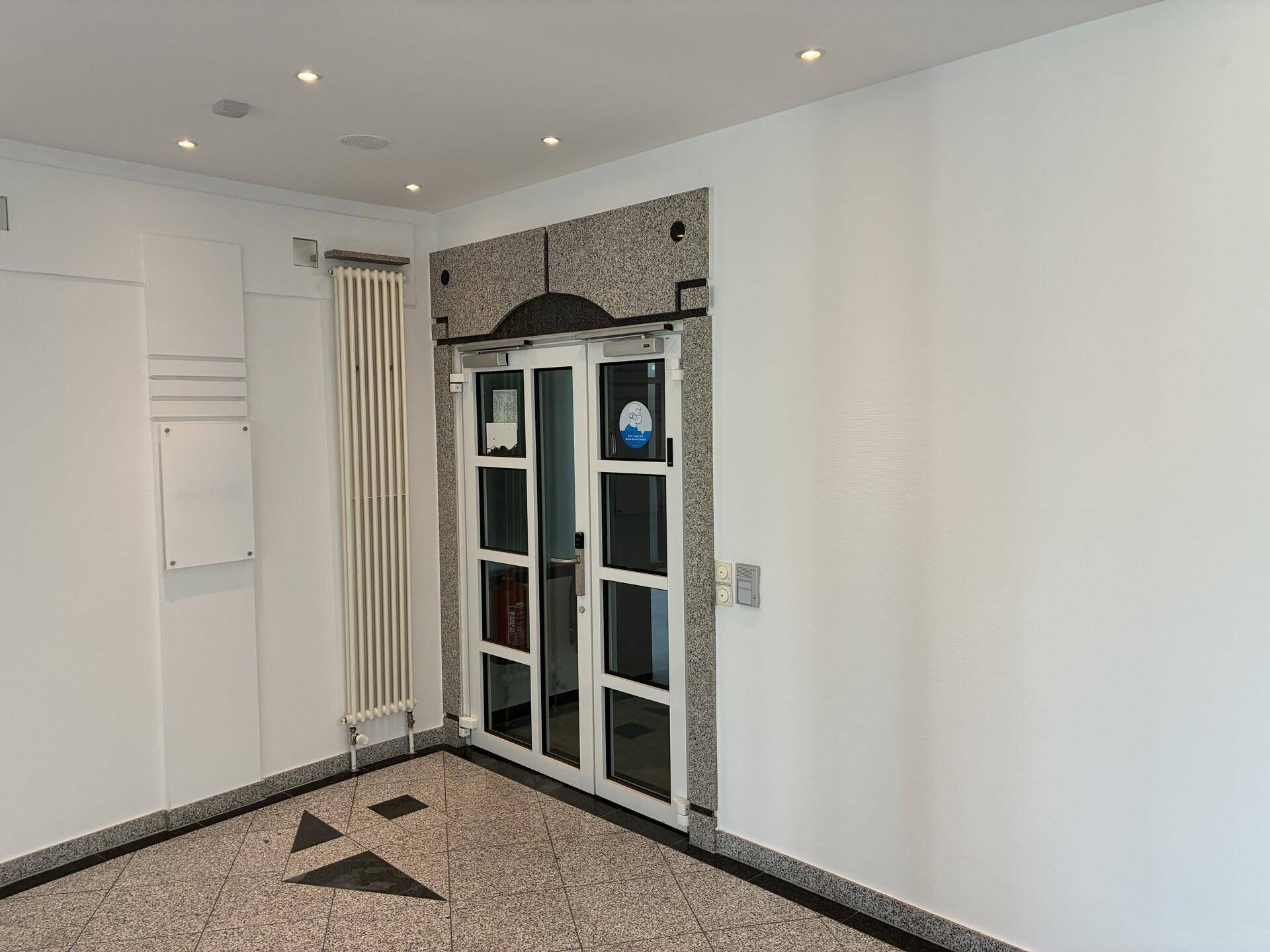 Eingangsbereich mit modernen Glastüren, Granitrahmen und einem hohen Heizkörper an der weißen Wand.
