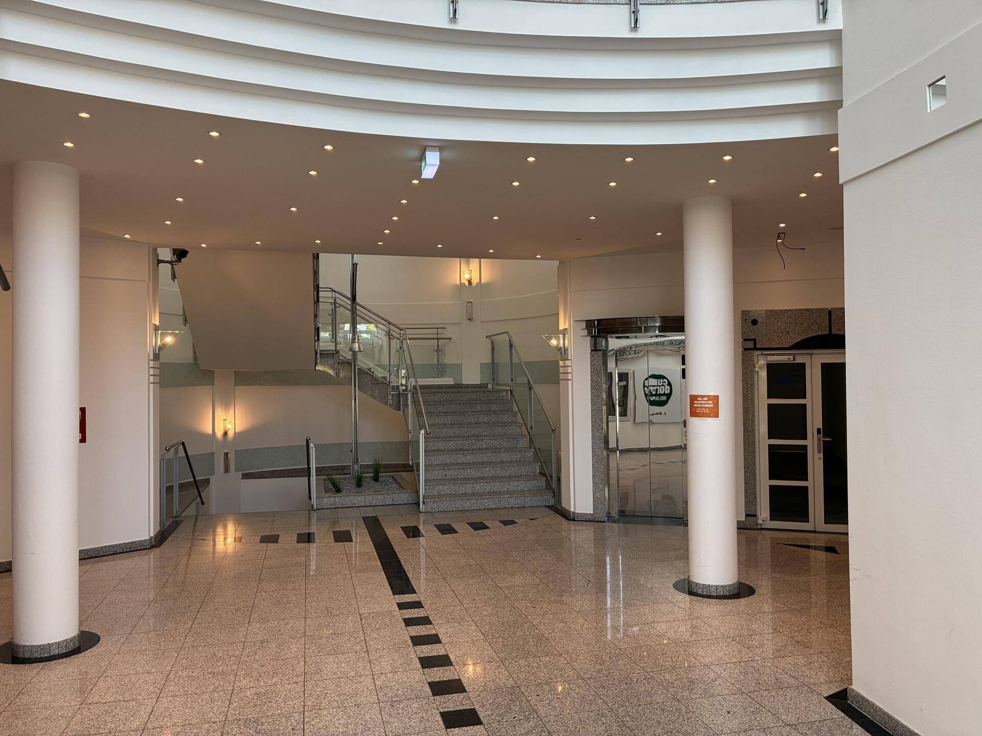 Moderner Eingangsbereich mit geschwungener Treppe, Glasgeländer und Säulen, die eine offene Atmosphäre schaffen.