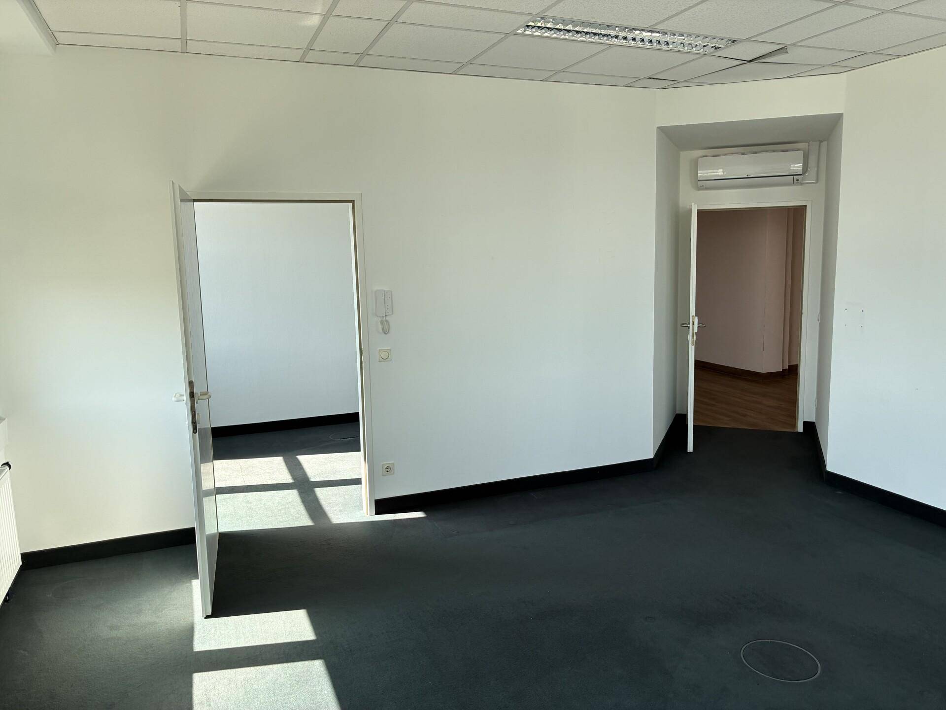 Leerer Büroraum mit dunklem Teppichboden, weißen Wänden und Klimaanlage über einer Tür.