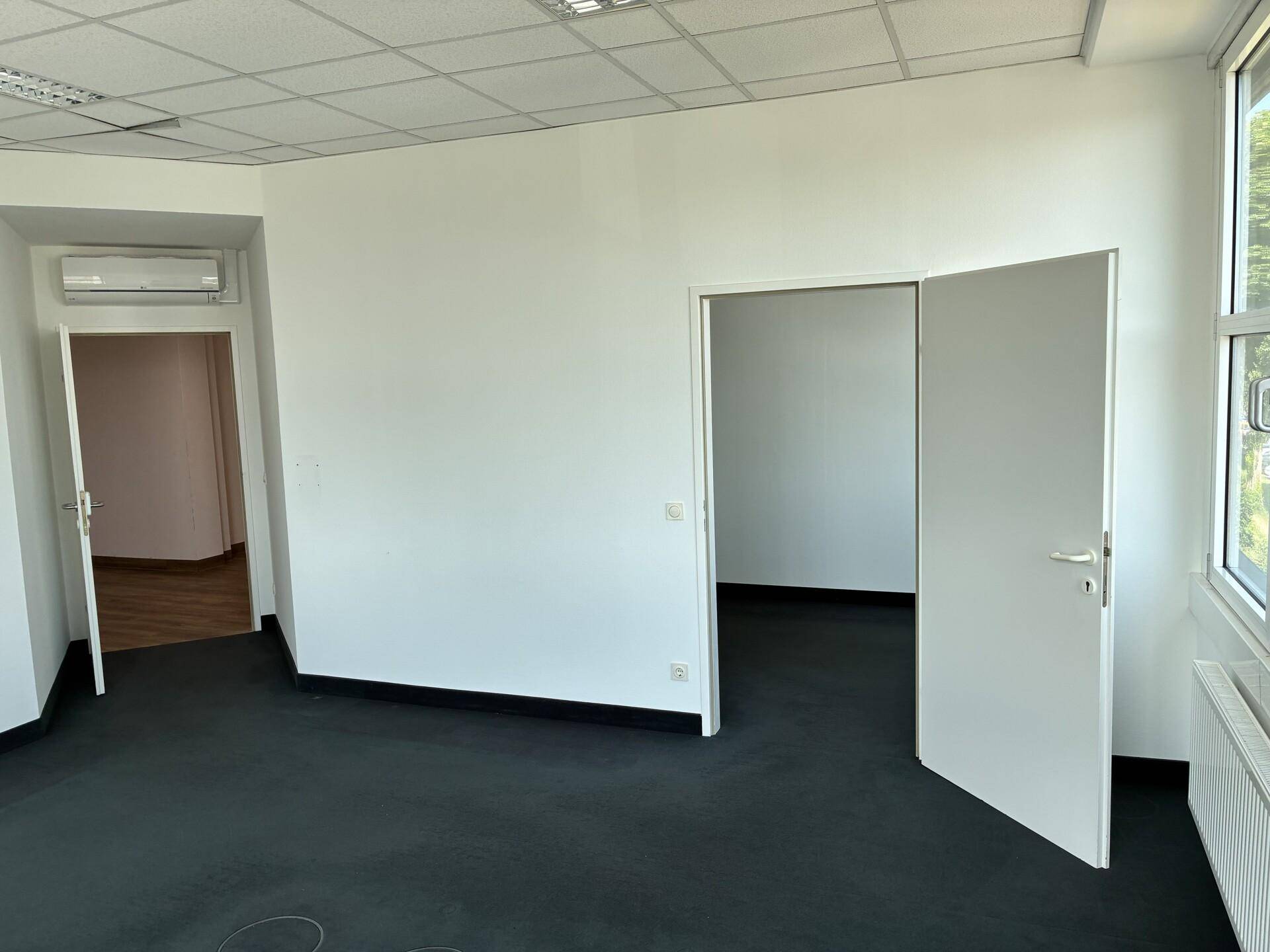 Geräumiger, leerer Büroraum mit dunklem Teppichboden, weißen Wänden und Klimaanlage.