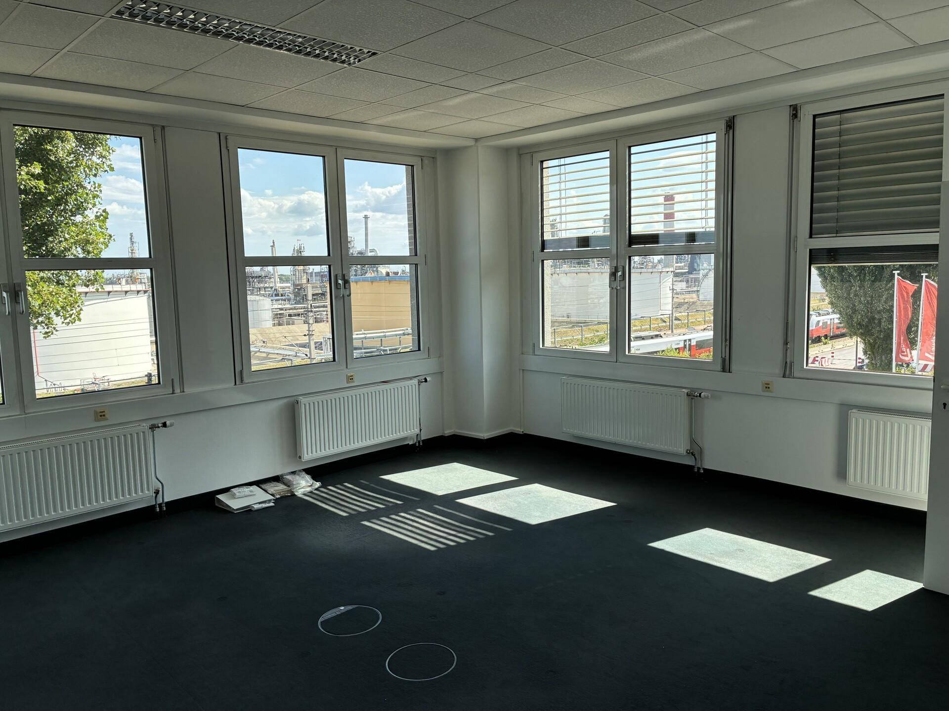 Heller Büroraum mit mehreren Fenstern, die einen Blick auf eine Industrieanlage bieten.