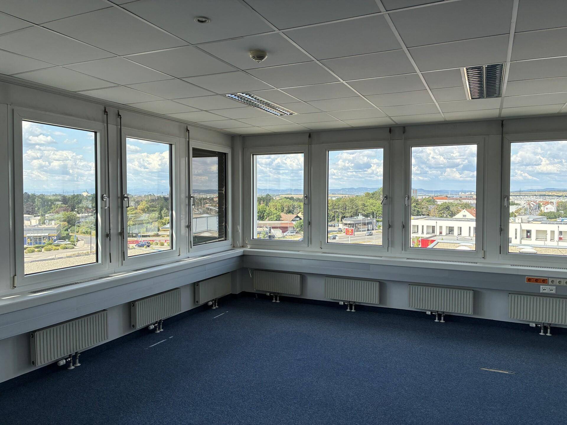 Eckbüro mit weitem Ausblick durch mehrere Fenster, blauer Teppichboden und Rasterdeckenbeleuchtung.