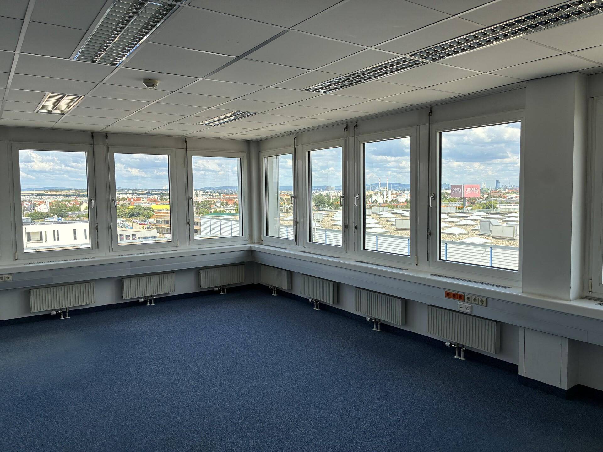 Geräumiges Eckbüro mit blauer Teppichboden, vielen Fenstern und einem Panoramablick auf die Umgebung.