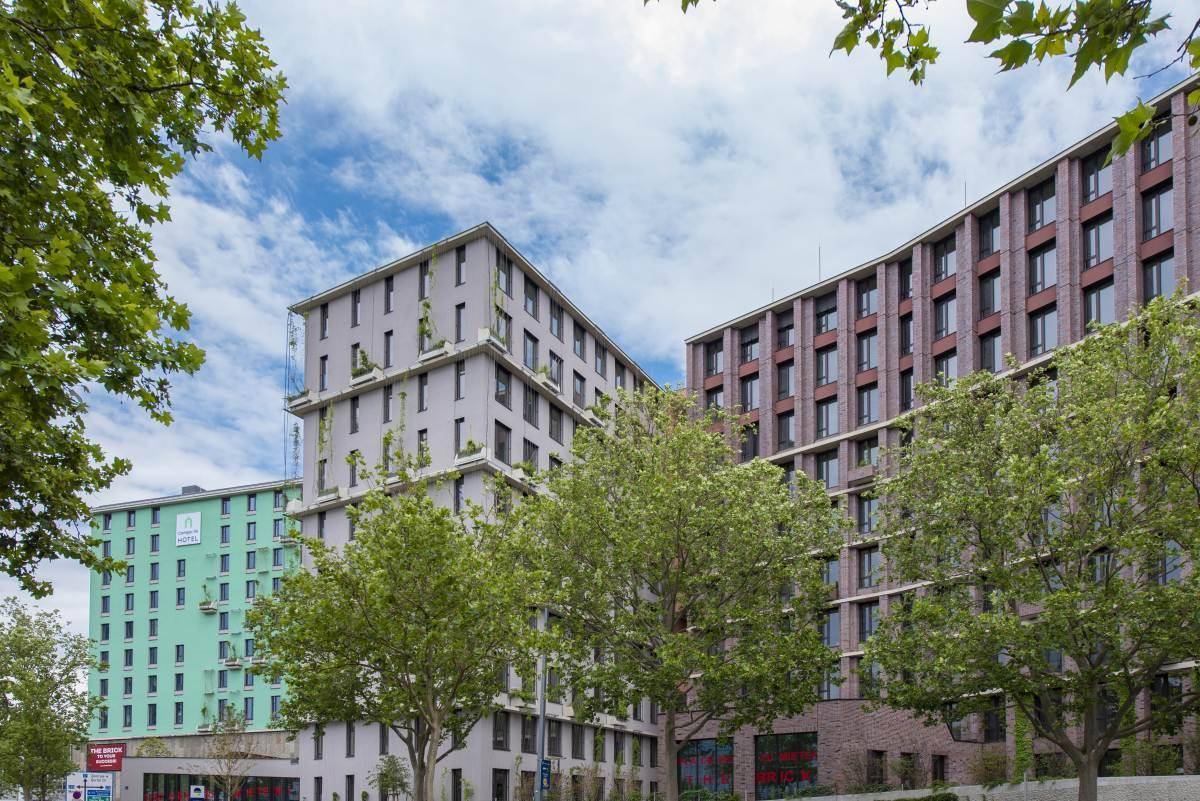 Ansicht eines modernen Gebäudekomplexes mit markanten grünen und ziegelroten Fassadenelementen an einer Straße.