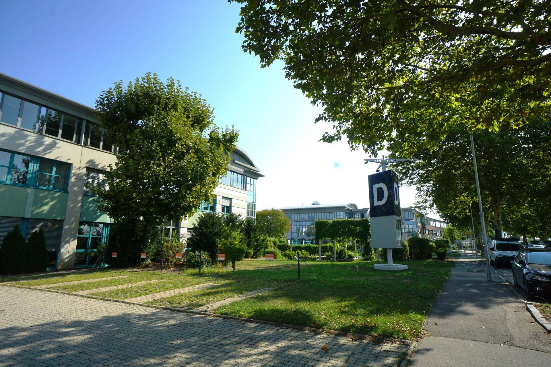Außenbereich des Bürokomplexes mit Grünflächen, Bäumen und einem Hinweisschild unter strahlendem Himmel.