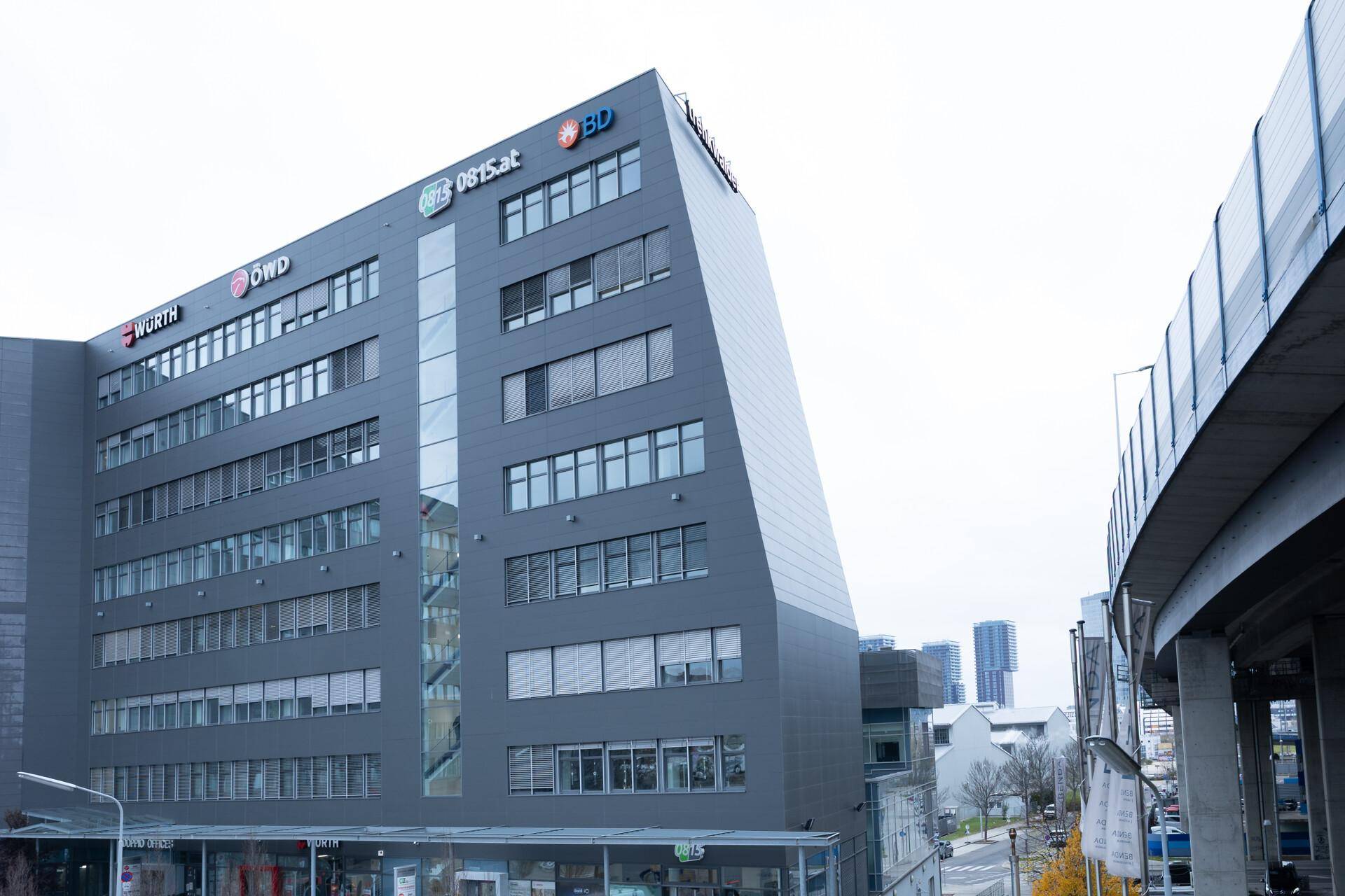 Weitwinkelaufnahme des modernen Bürogebäudes mit markanter Architektur und einer Überführung im Vordergrund, die die städtische Umgebung betont.