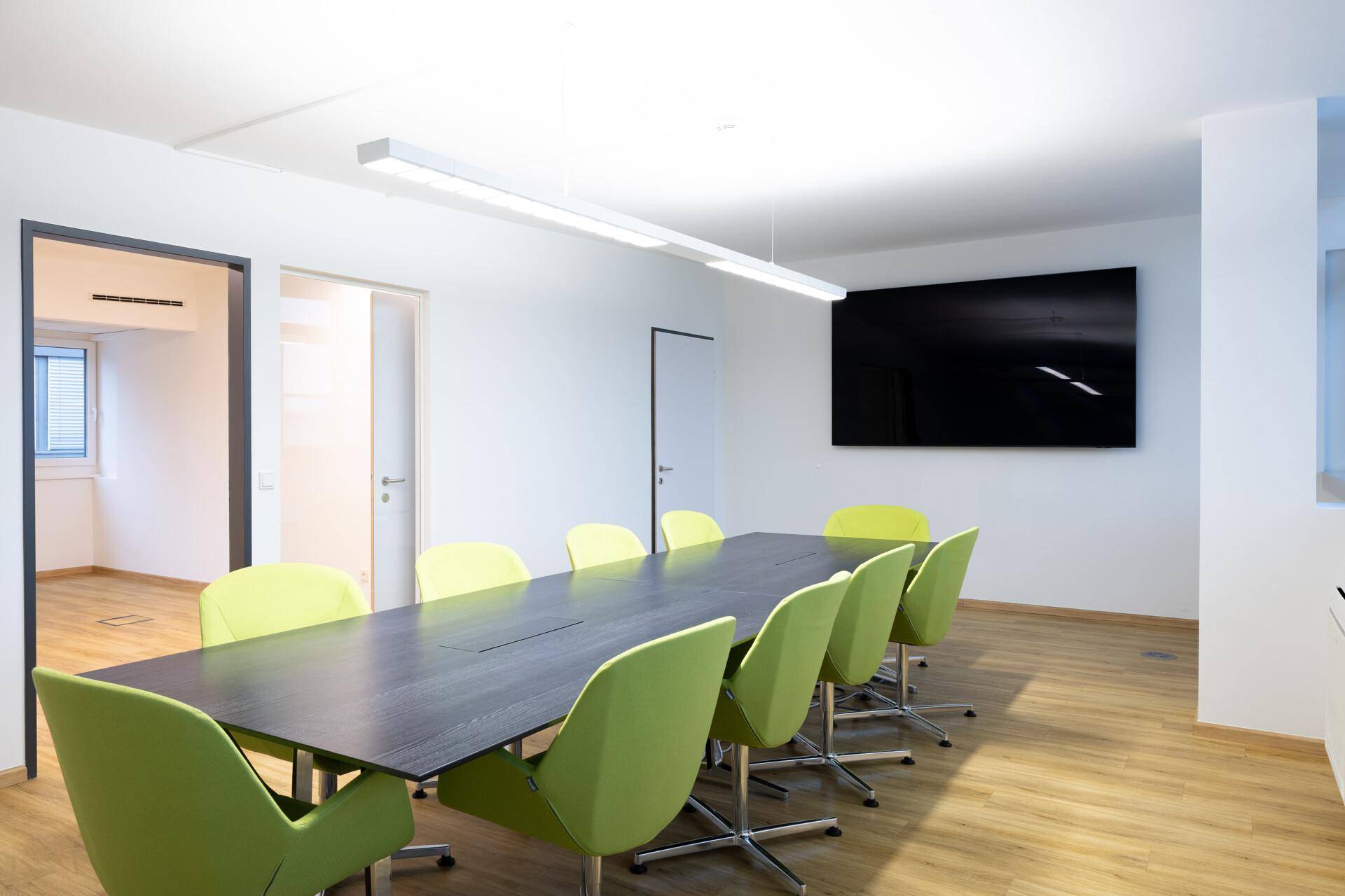Konferenzraum mit langem dunklem Tisch, leuchtend grünen Stühlen und einem großen Wandbildschirm für Präsentationen, sehr modern eingerichtet.