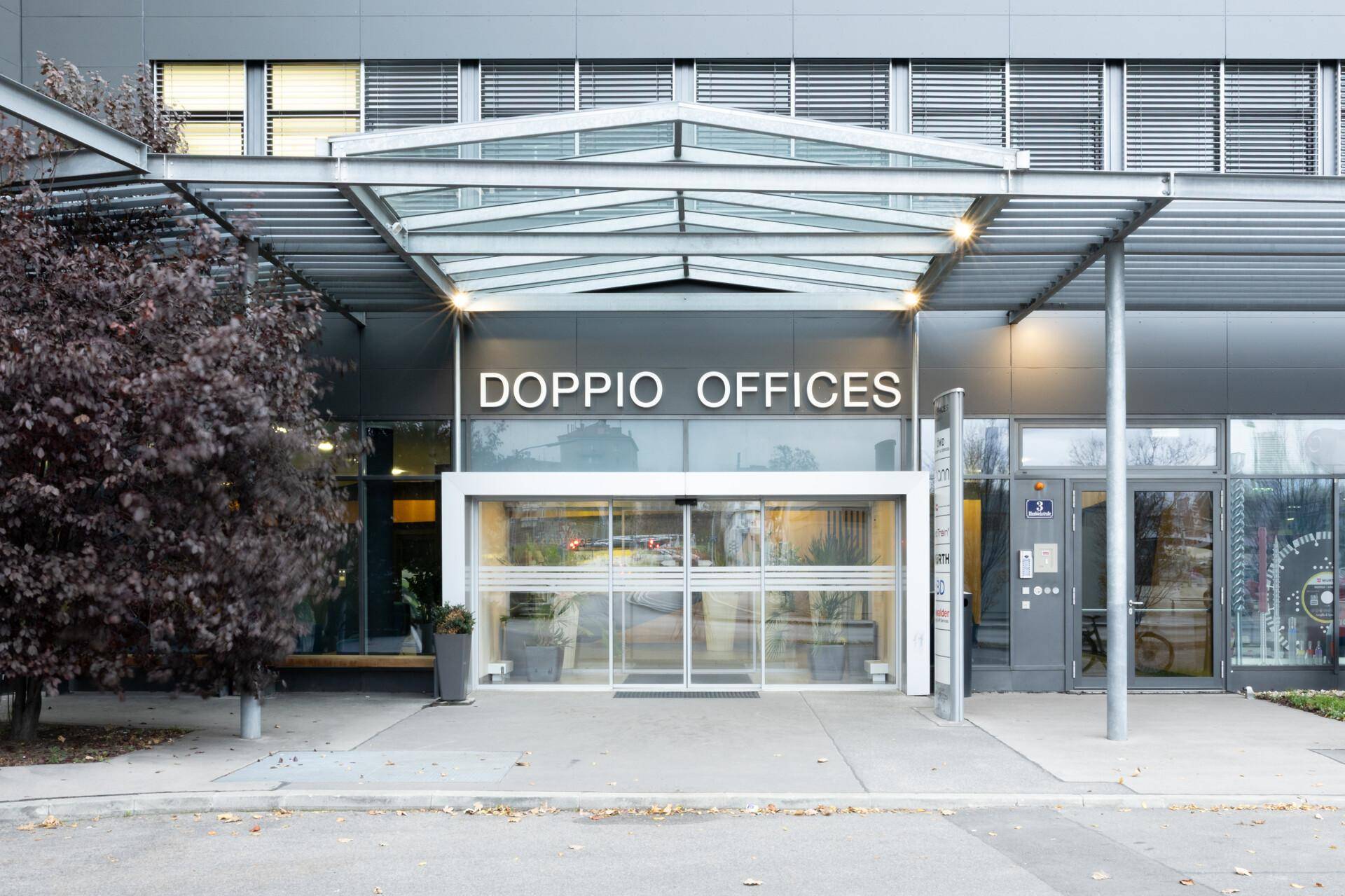 Eingangsbereich der Doppio Offices mit Glasfassade und einem schützenden Vordach, umgeben von Bäumen, was einen einladenden Eindruck vermittelt.