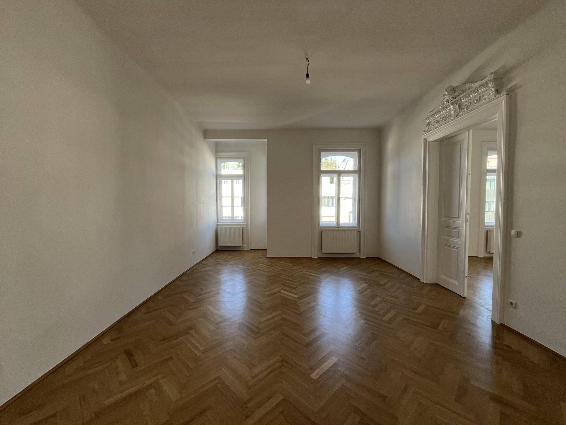 Großes, leeres Zimmer mit Fischgrätparkett und hohen Decken, ideal für ein Wohnzimmer.