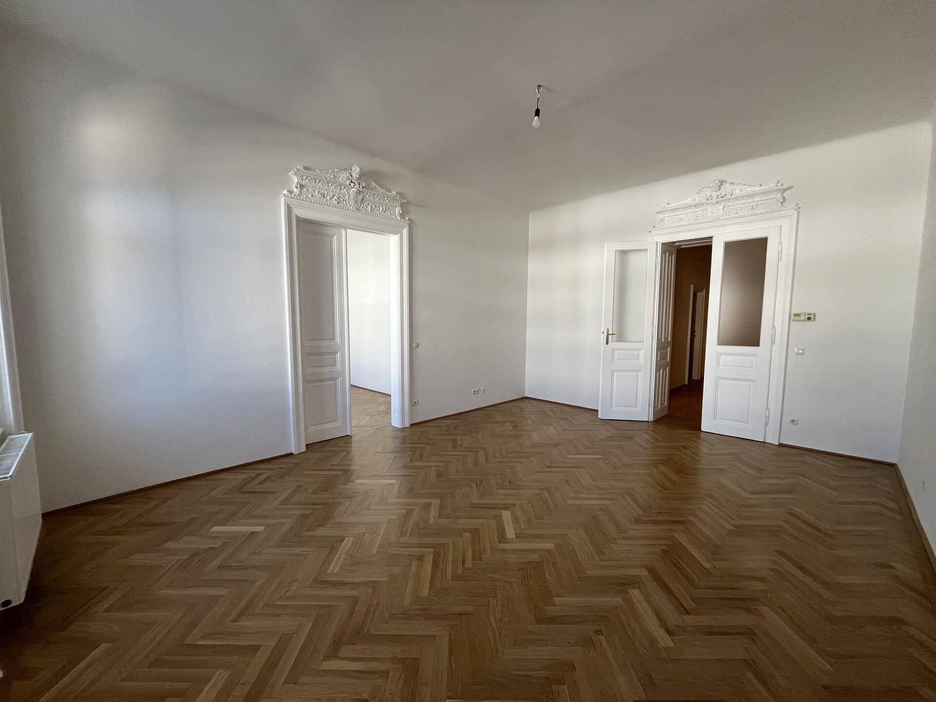 Geräumiges Wohnzimmer mit Fischgrätparkett und dekorativen Türrahmen, bereit zur individuellen Gestaltung.