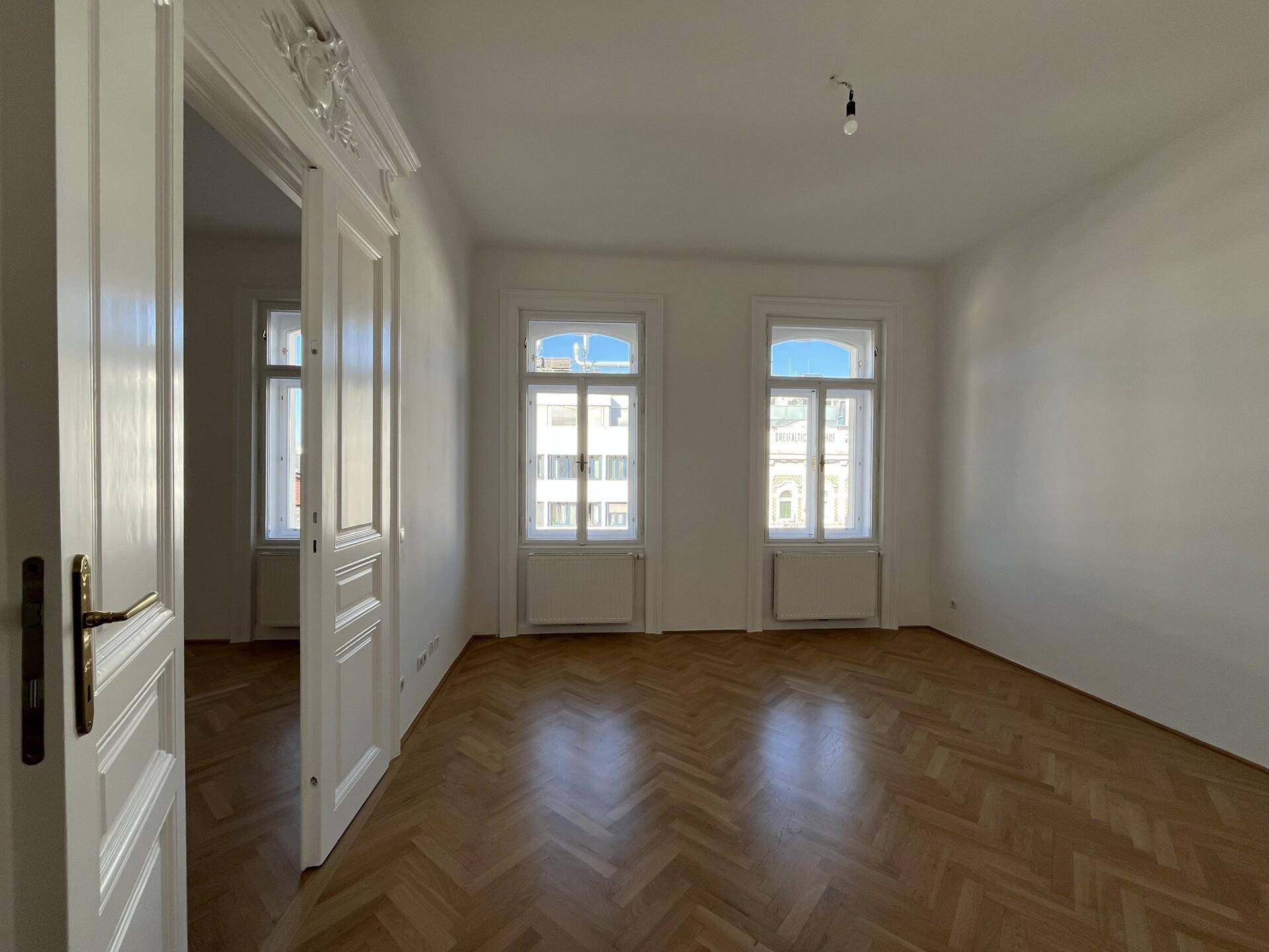 Helles Wohnzimmer mit Fischgrätparkett und hohen Fenstern, die viel Tageslicht hereinlassen.