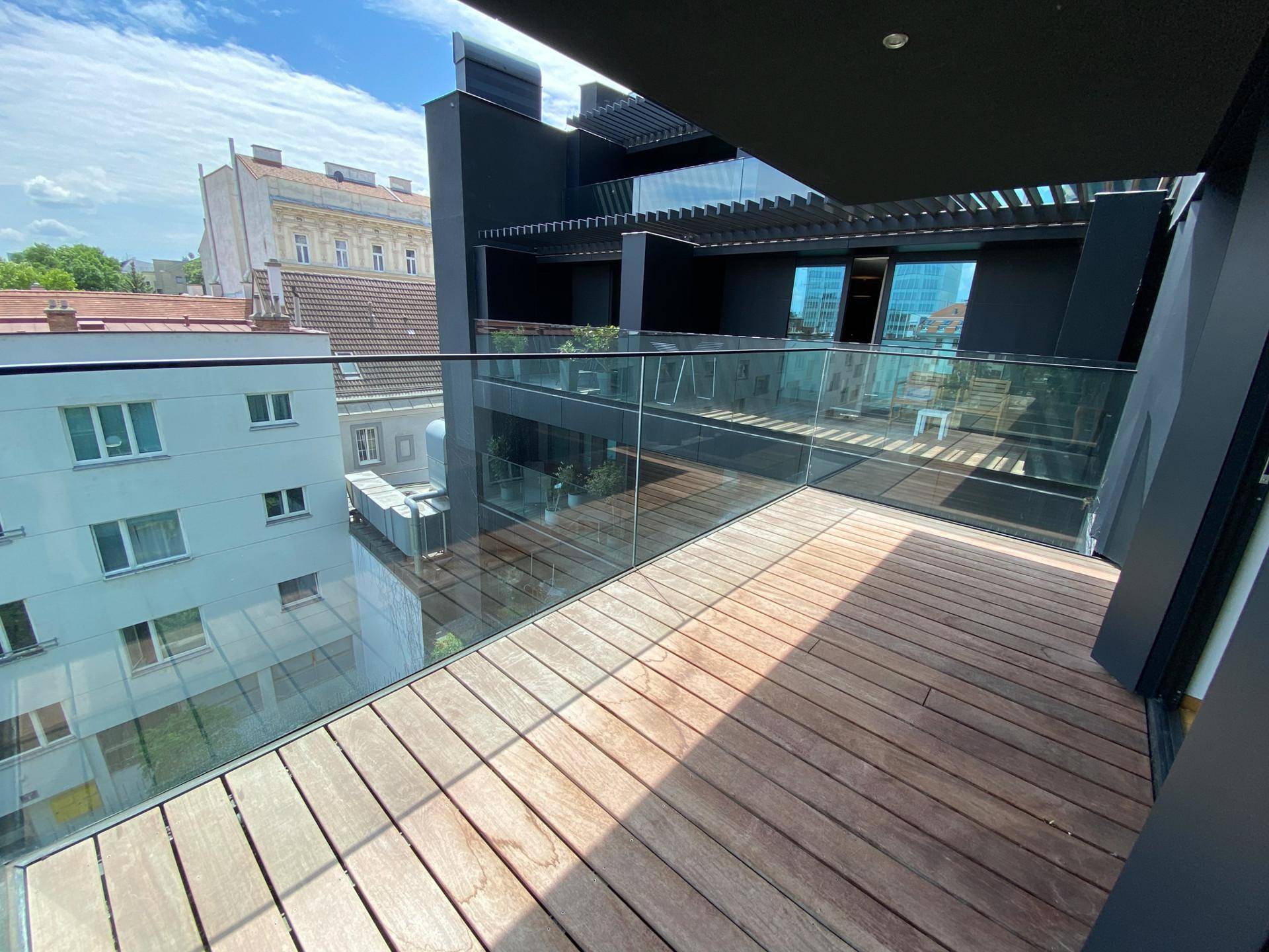 Moderner Balkon mit Holzboden, Glasgeländer und Blick auf die umliegende Architektur.