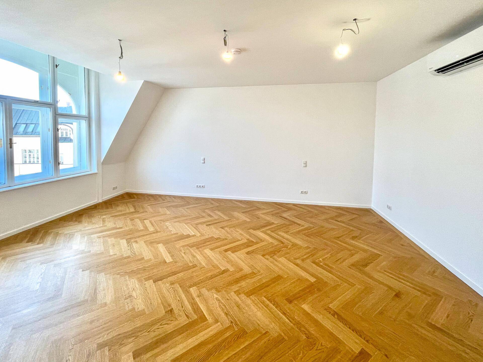 Lichtdurchflutetes Zimmer mit Fischgrätparkett, Dachschrägen und einem großen Fenster, ausgestattet mit Klimaanlage.