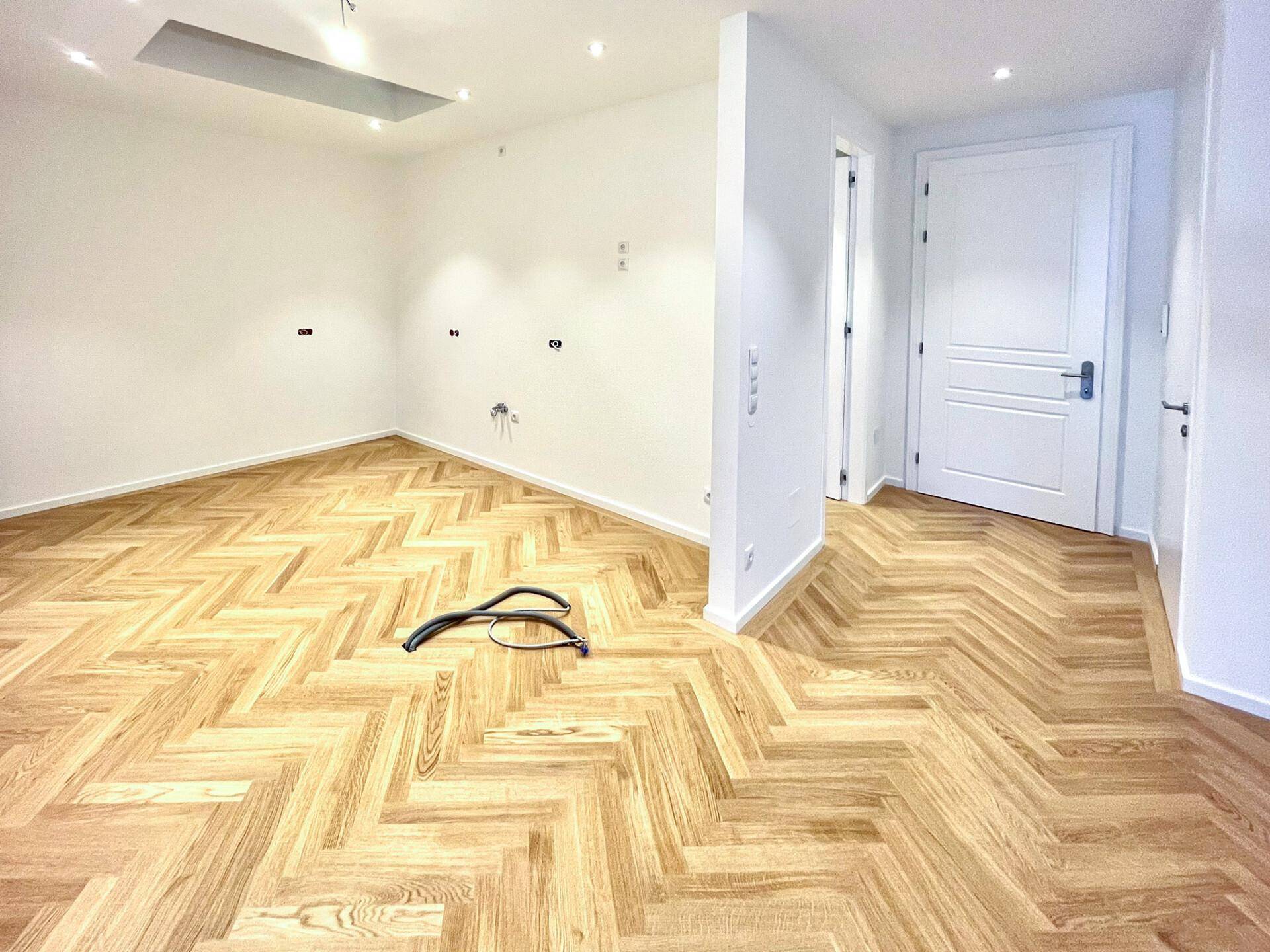 Leerer Küchenbereich mit Fischgrätparkett, vorbereiteten Anschlüssen und Einbaustrahlern, bereit für die Installation.