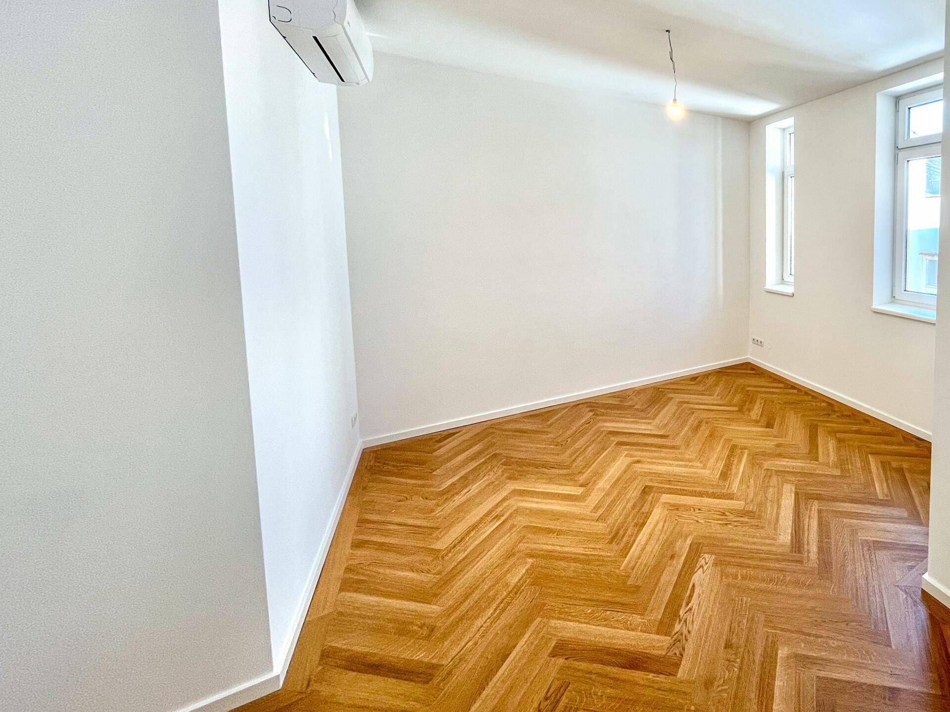 Helles Schlafzimmer mit Fischgrätparkett, weißen Wänden und zwei Fenstern, ausgestattet mit Klimaanlage.