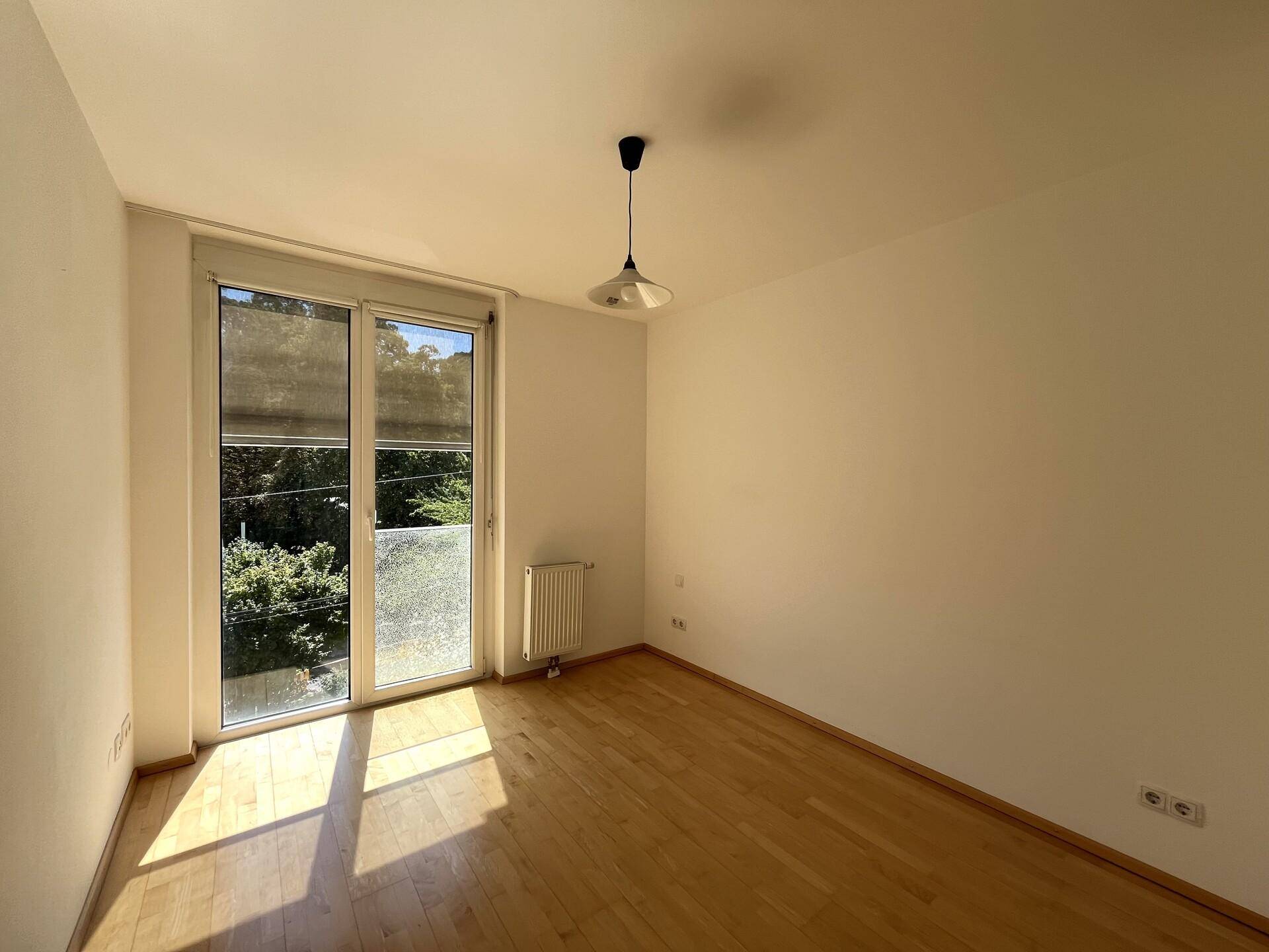 Helles Schlafzimmer mit Parkettboden und großem Fenster, das Zugang nach draußen bietet.
